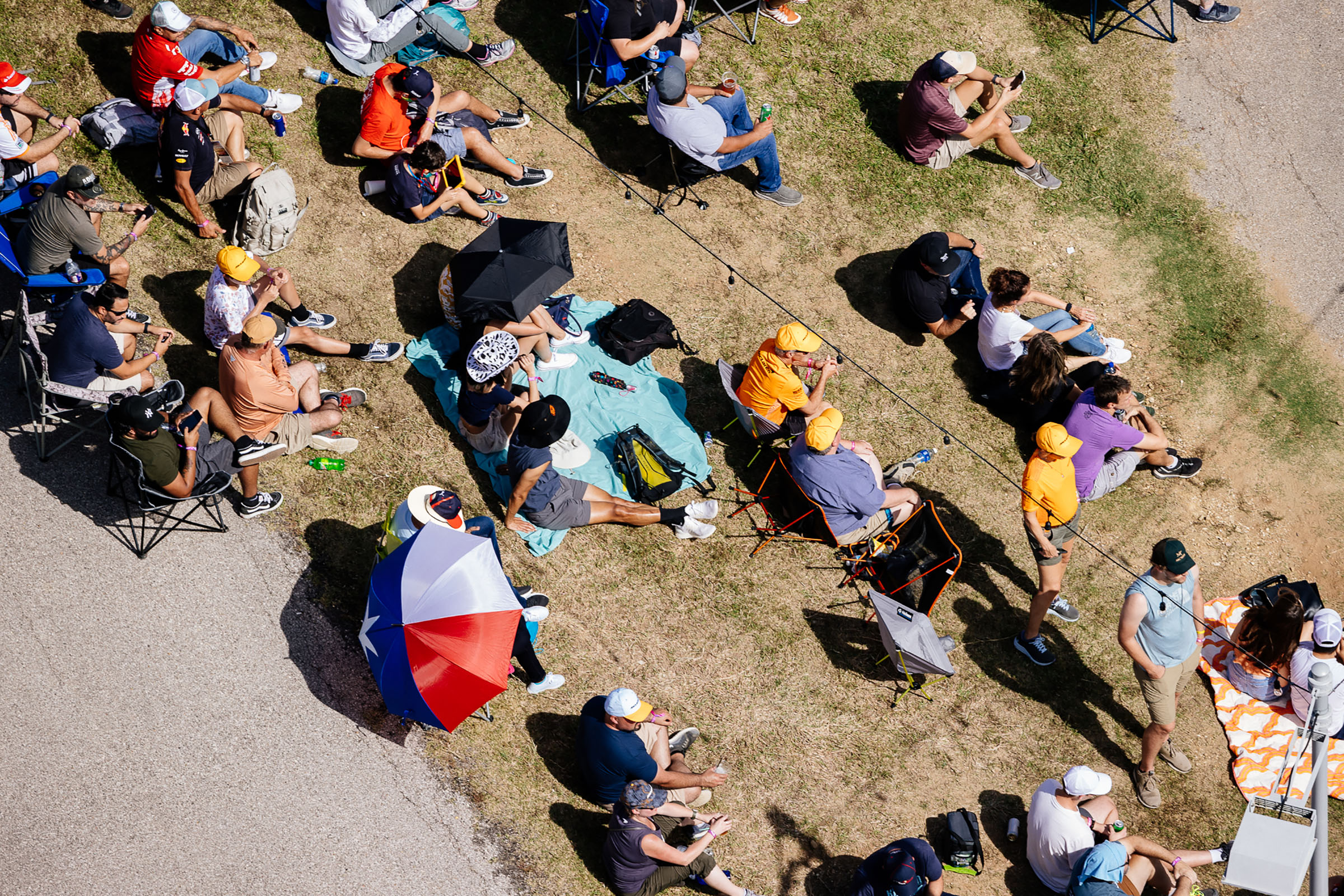 From above, a collection of people sitting on brown grass, some in lawn chairs, and several holding umbrellas to block the sun