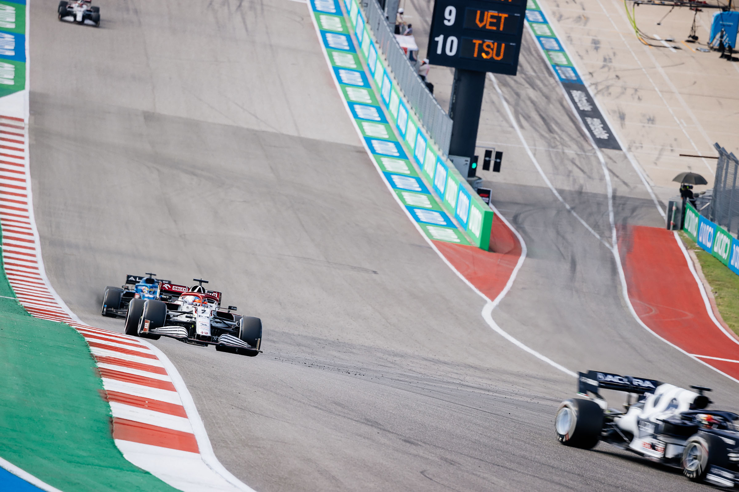 An overhead view of three cars moving up a gray, hilly track, with a scoreboard in the background