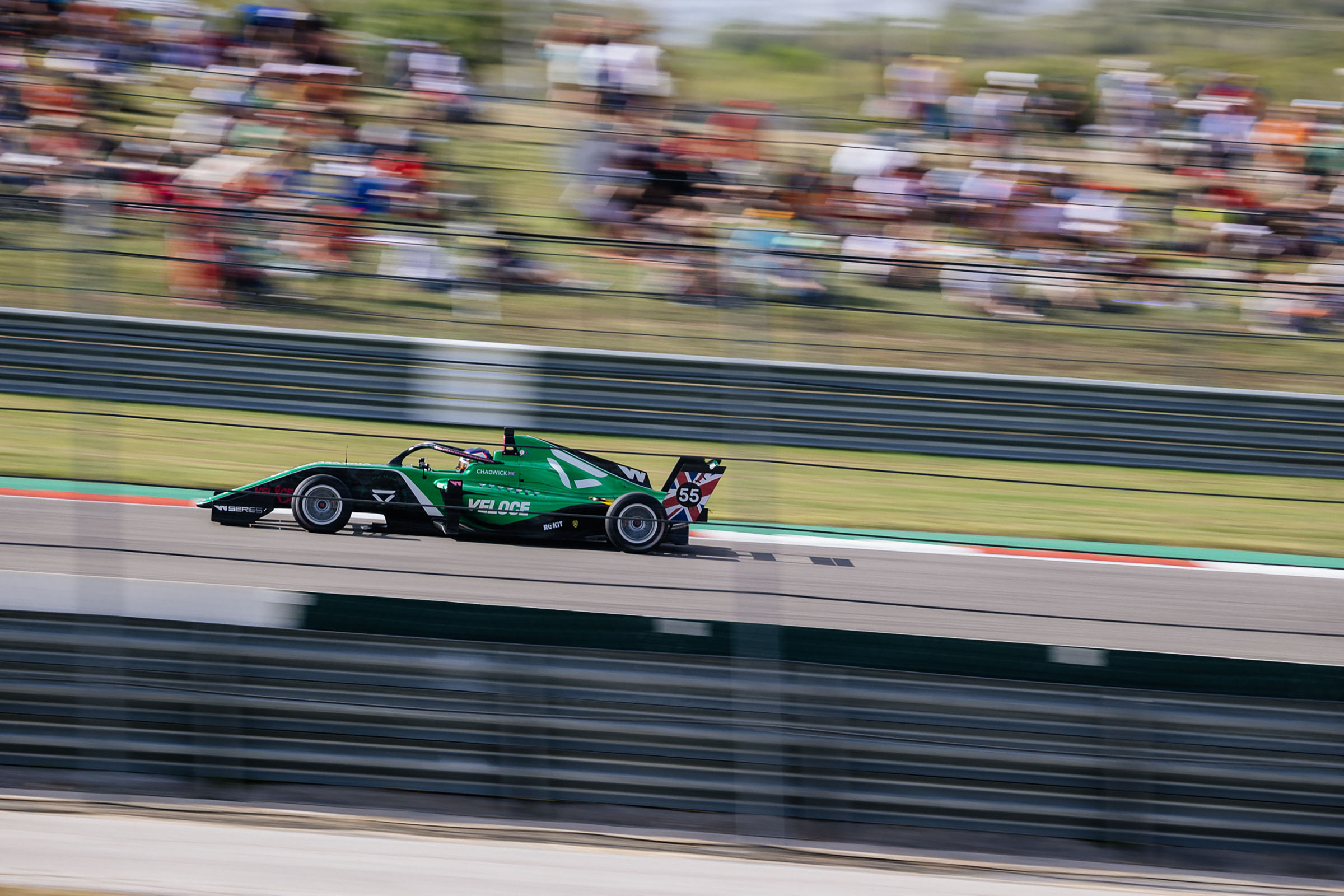 A side view of a green car racing down a track with blurred groups of people in the background