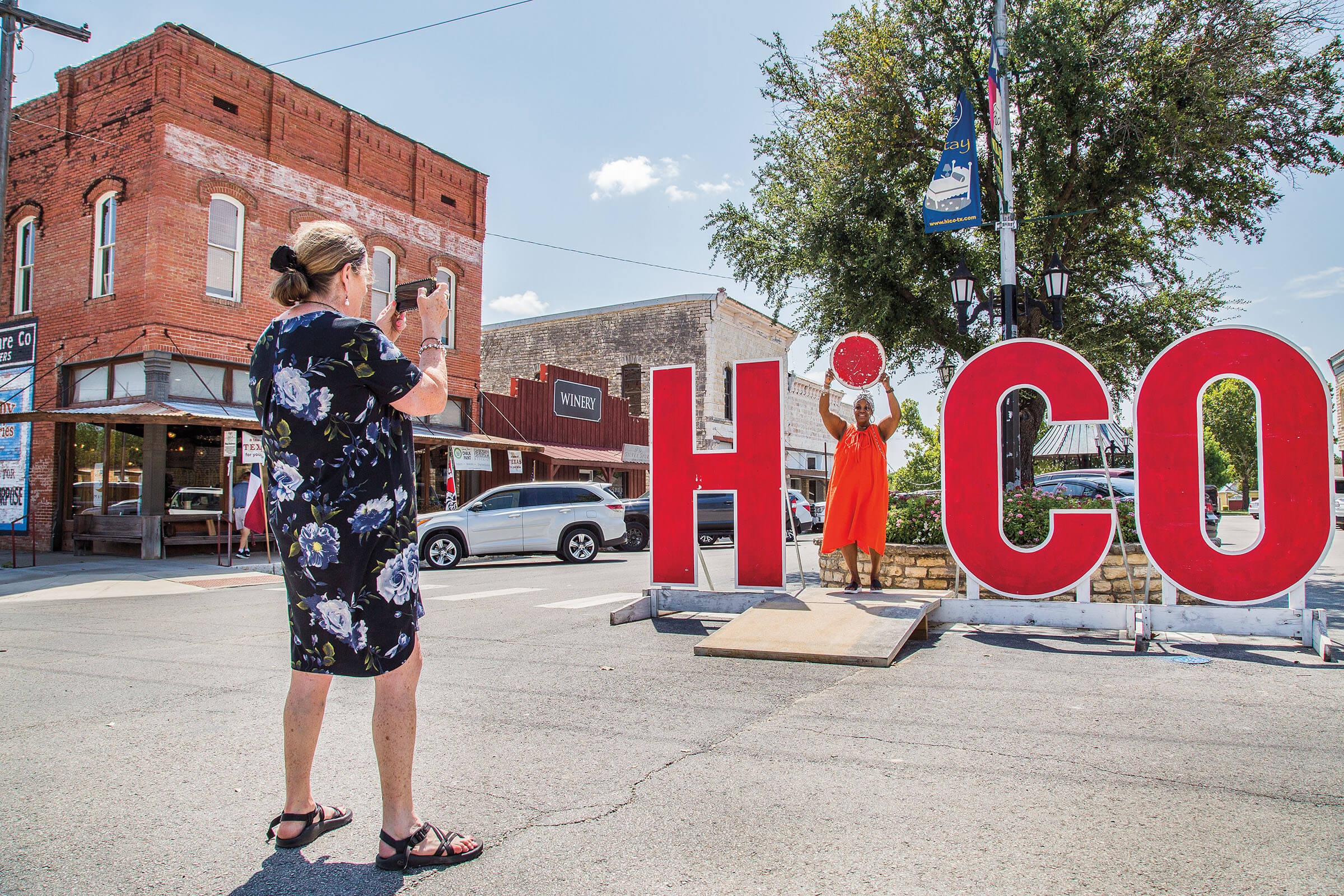 A woman in a black dress points a camera at her friend, who is standing inside bright red letters reading HICO