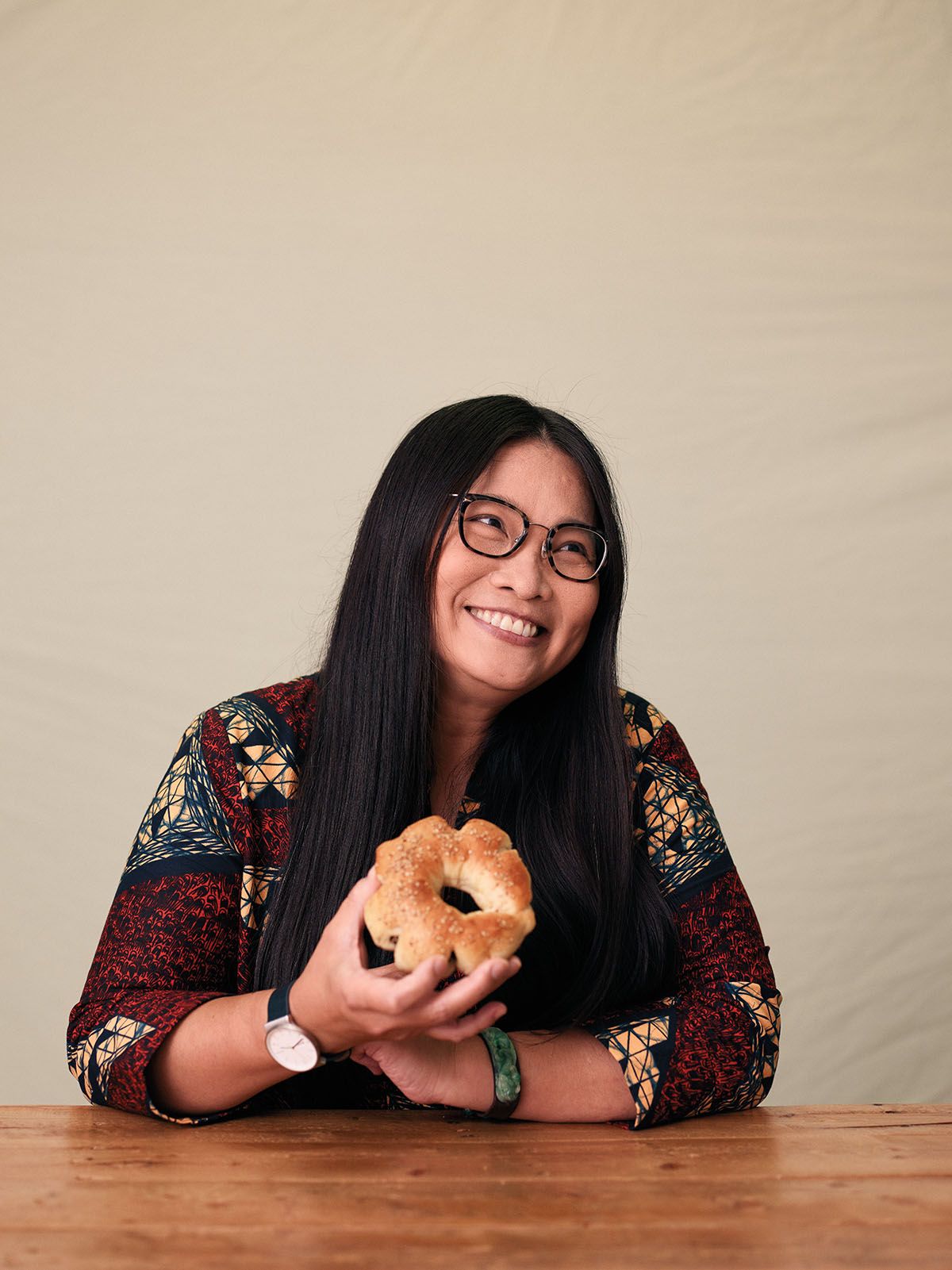 A woman with long dark hair and glasses smiles while holding a bagel in front of a plain background