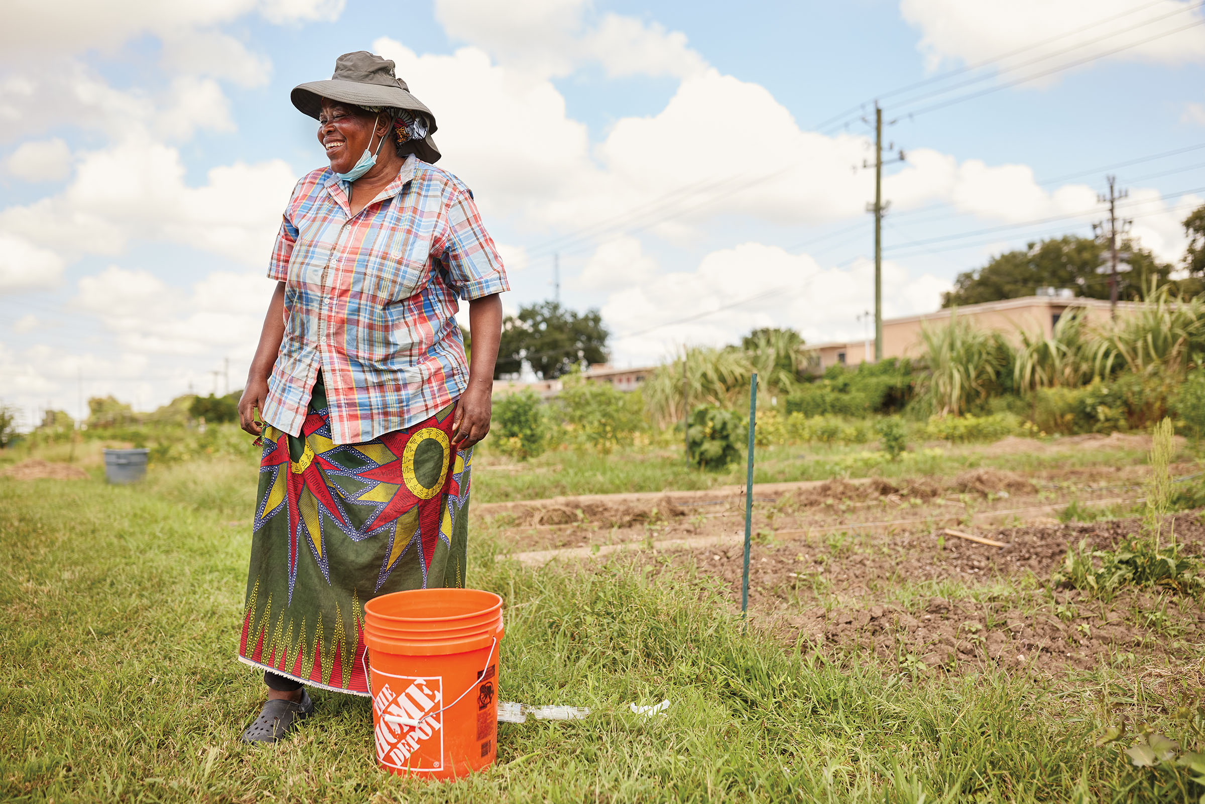 A woman in a hat stands in a field with an orange bucket under a blue sky in front of crops