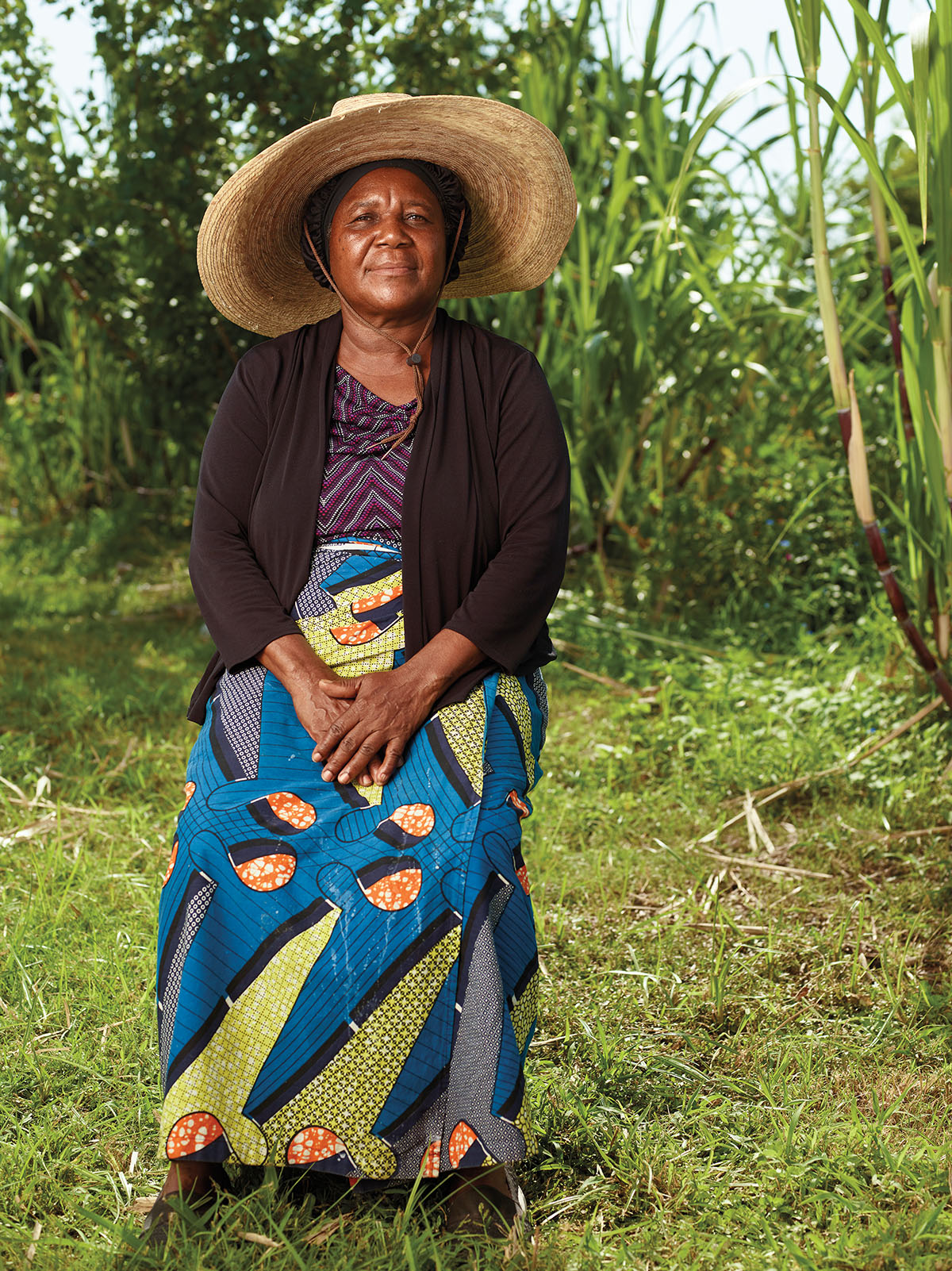 A woman in a black blazer, brightly colored skirt and straw hat looks into the camera in a green field