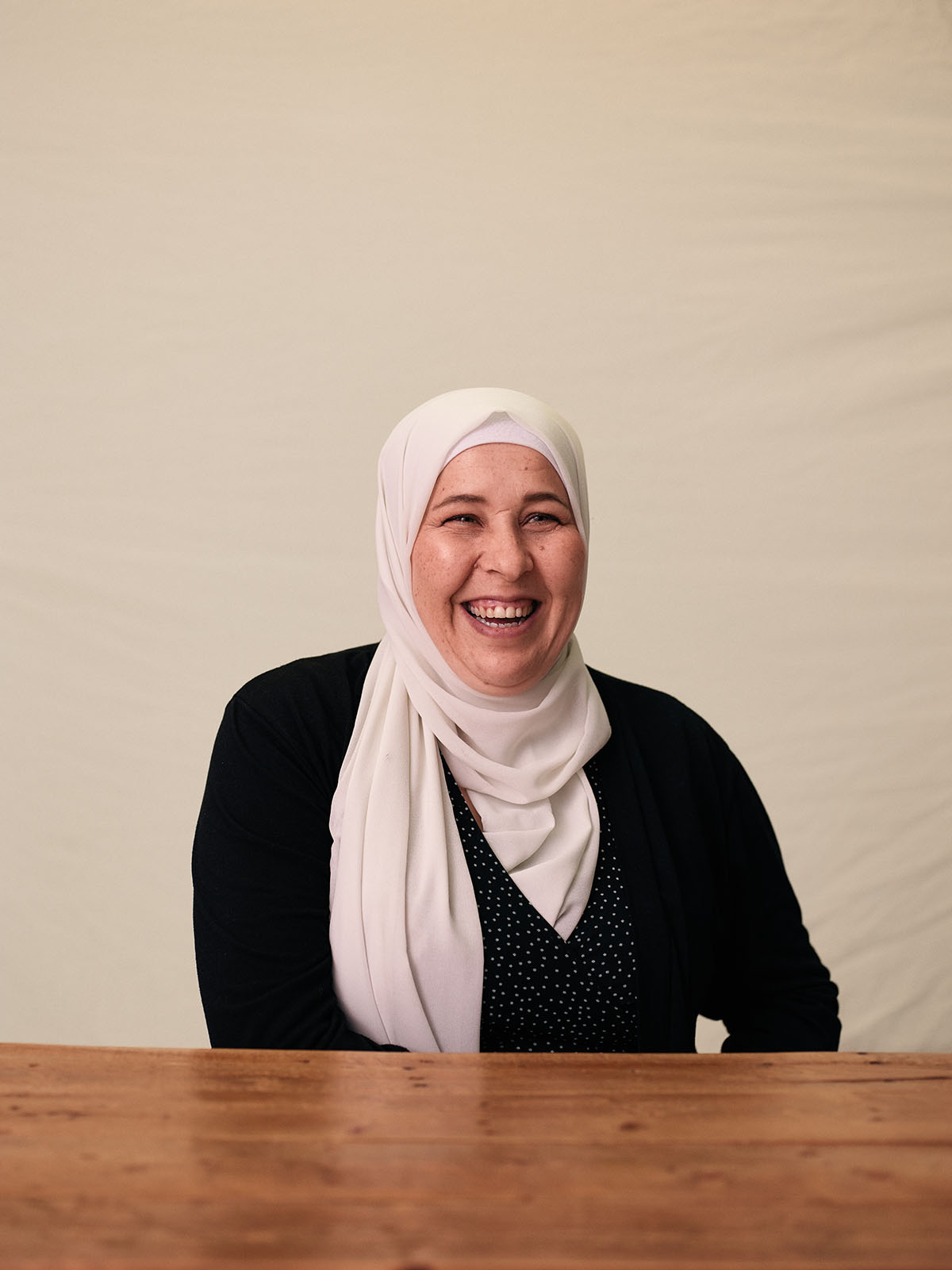 A woman in a white hijab and black shirt smiles in front of a plain background