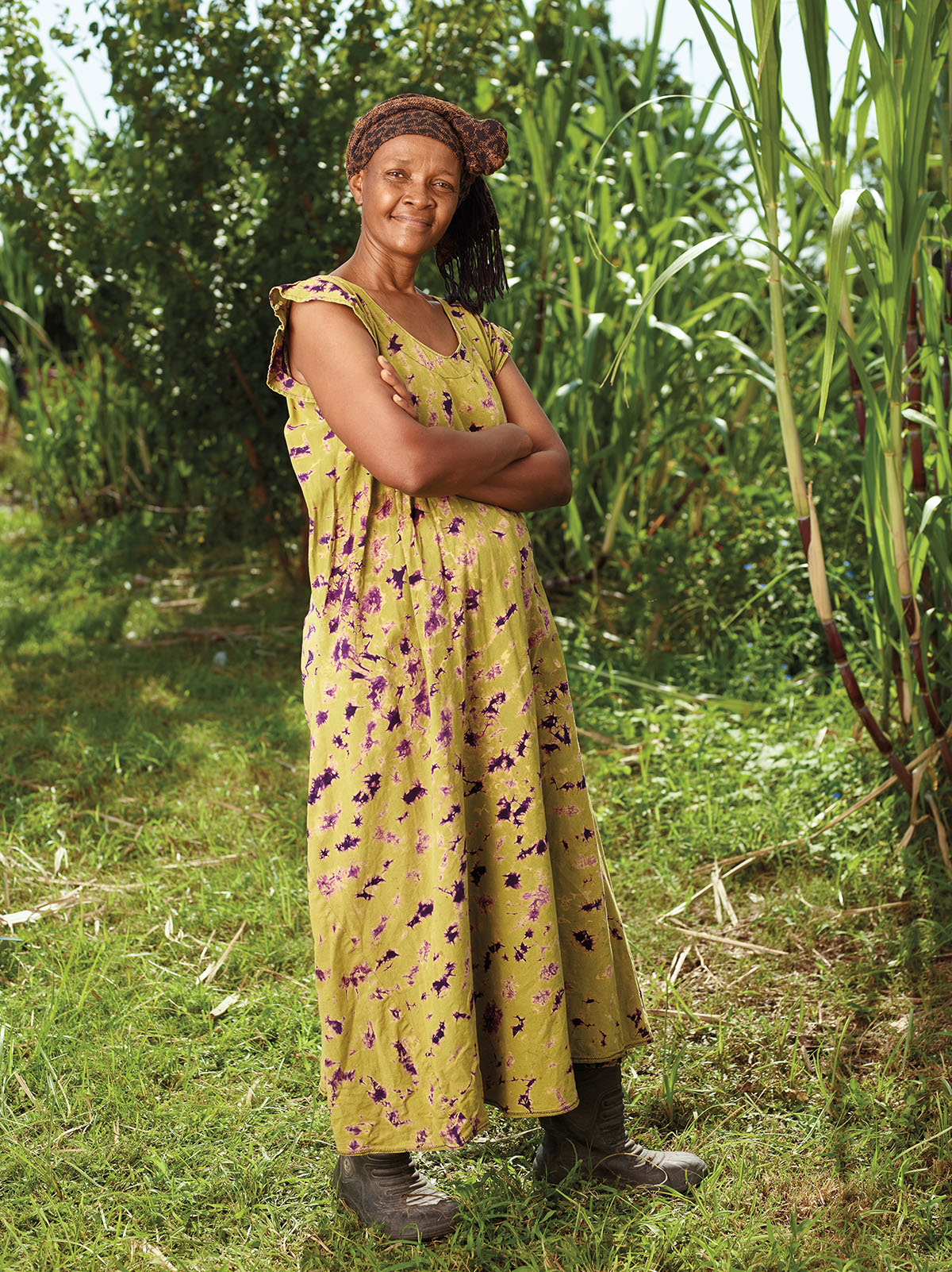 A woman in a yellow patterned dress stands with her hands folded in a green field