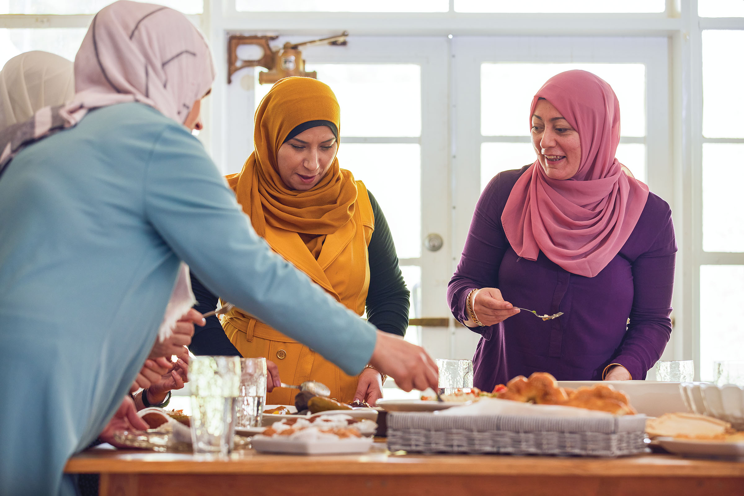 A group of women in brightly-colored hijabs talk in a kitchen around a plate of food