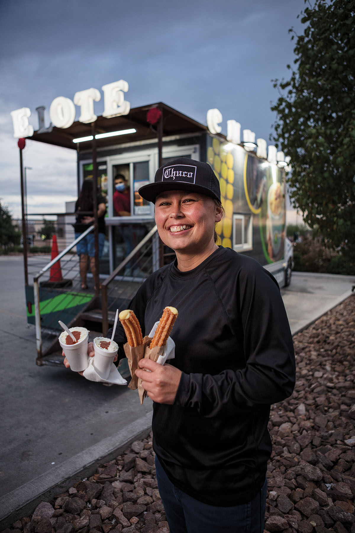 A person in a black flat-brimmed ball cap stands with two cups of elotes in one hand and two churros in the other