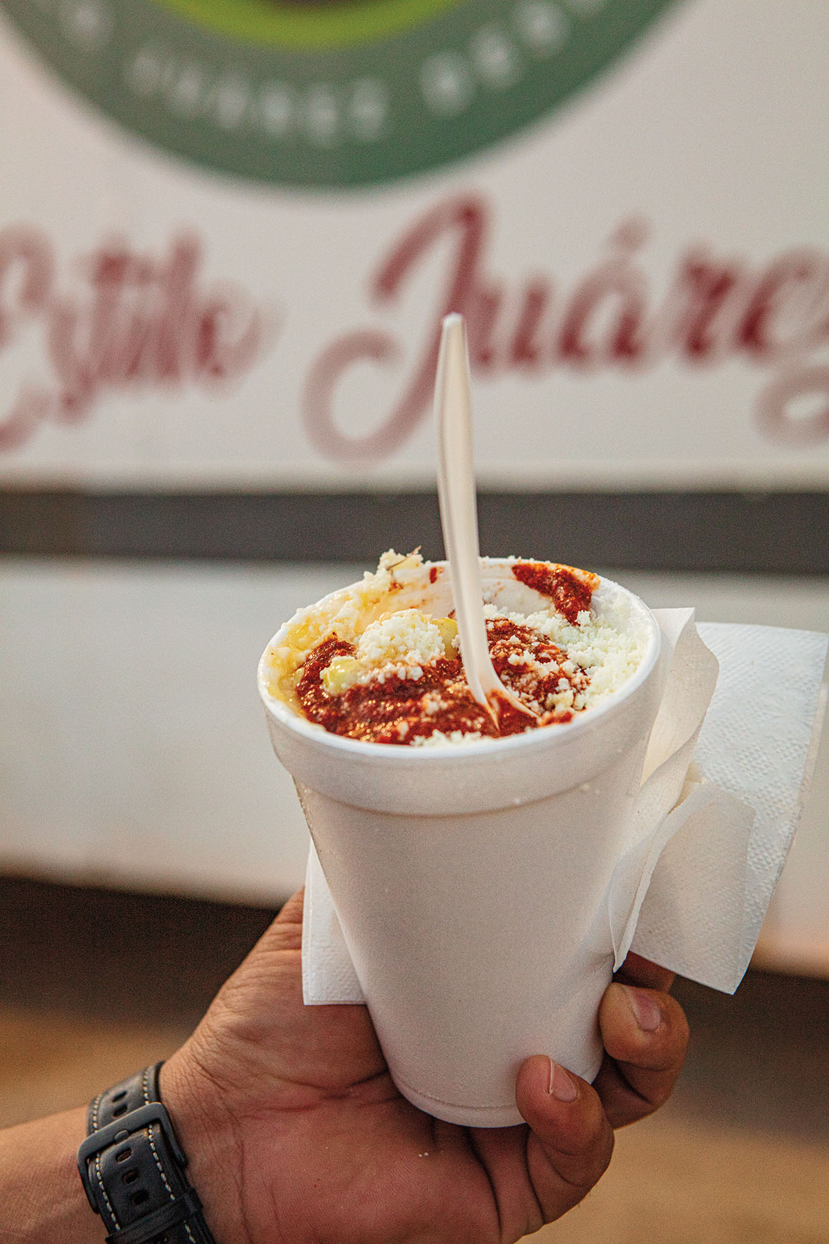 A hand holds a styrofoam cup filled full of corn, topped with red chile powder, cheese and a plastic spoon