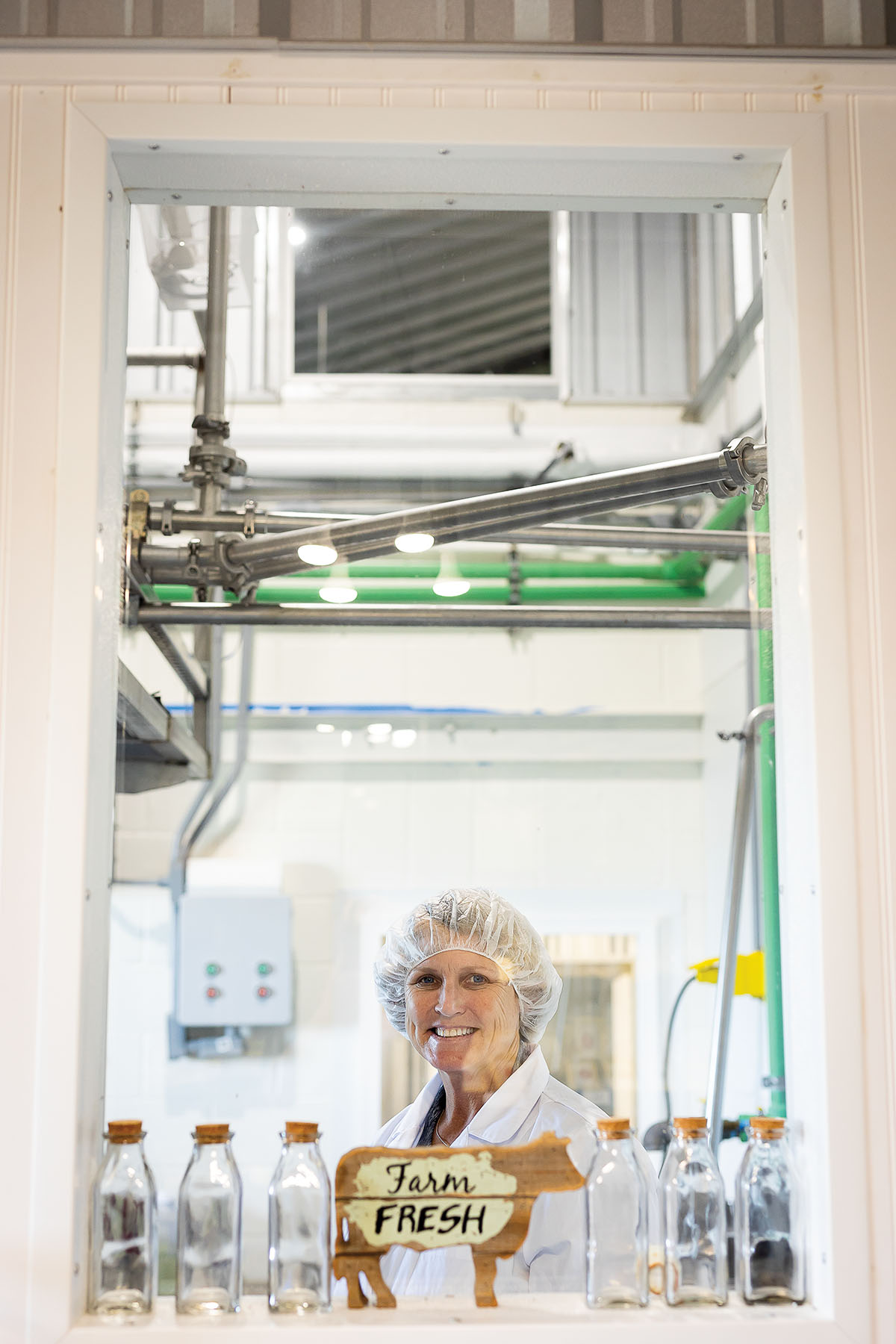 A woman in a white hair net stands behind empty glass milk bottles and a sign reading "Farm Fresh"