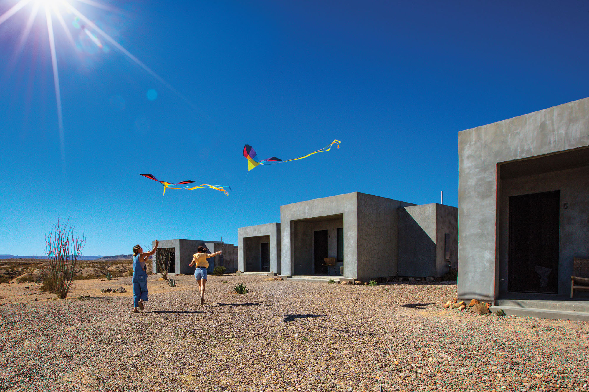 Large gray concrete boxes along the right side frame two people flying colorful kites in the bright blue sky above a brown desert scene