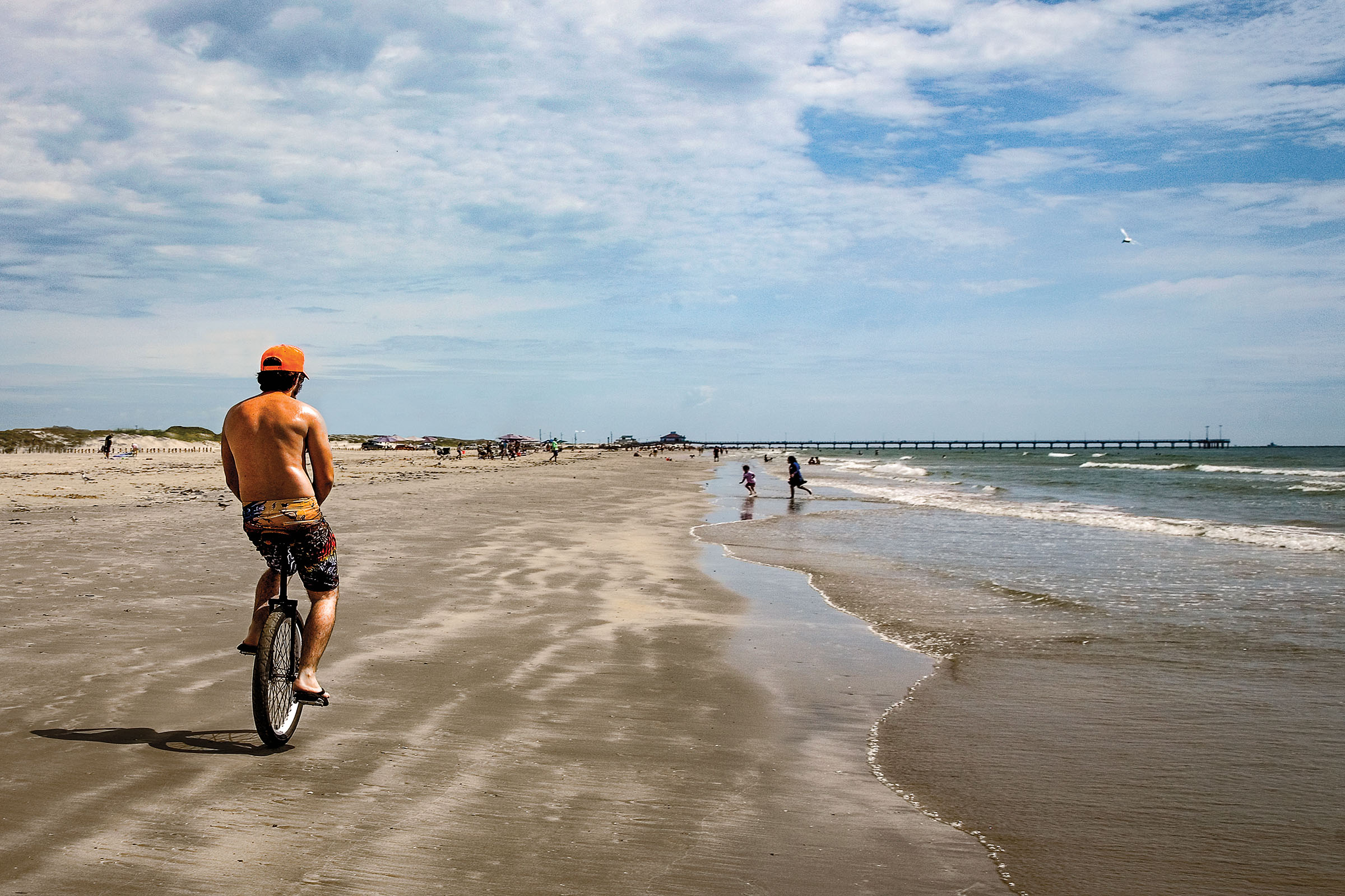 A unicyclist pedals along the hard-packed sand along the seashore under blue sky