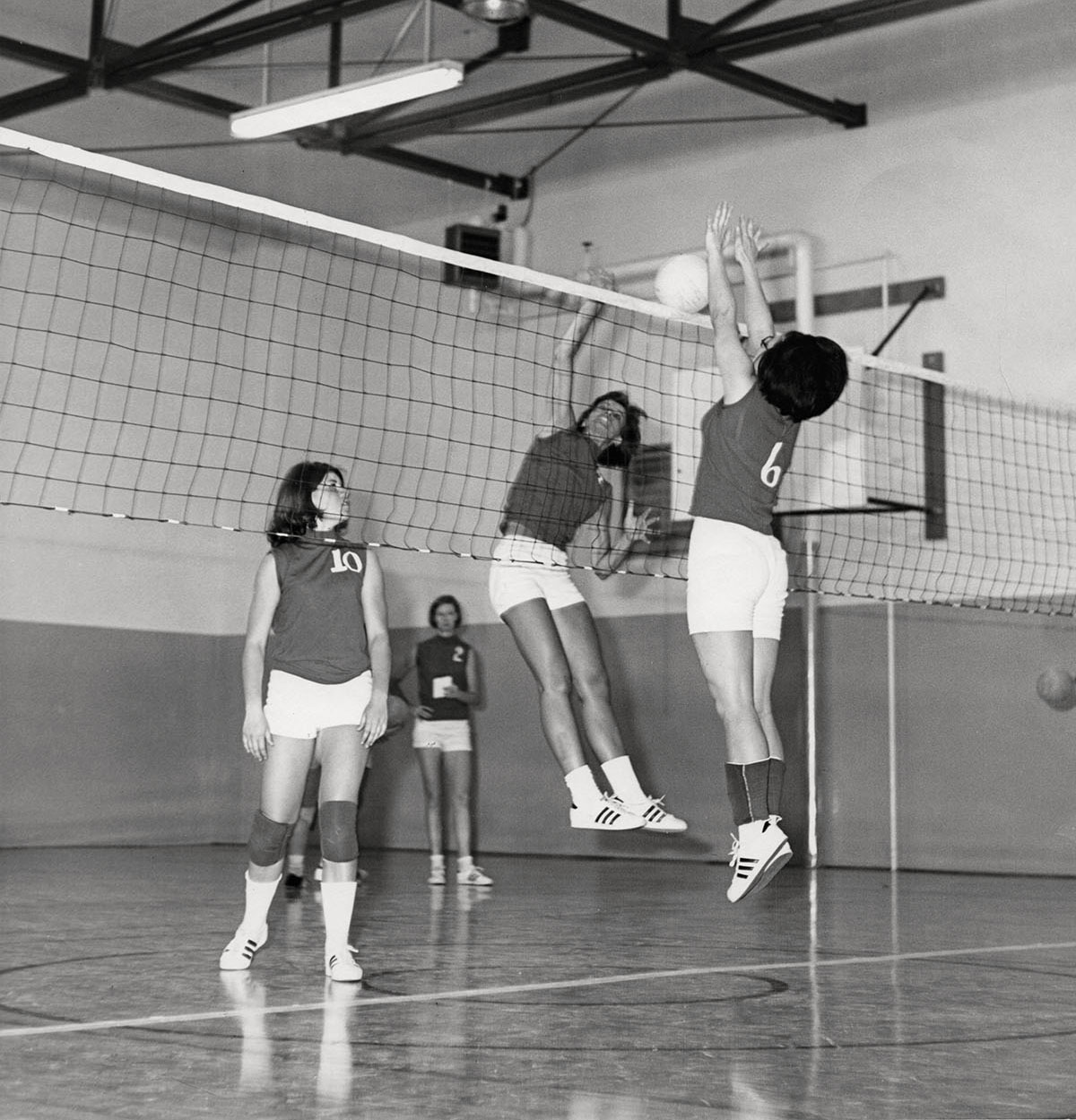 One woman reaches up toward a net while another puts two hands in the air to block a volleyball in a gymnasium