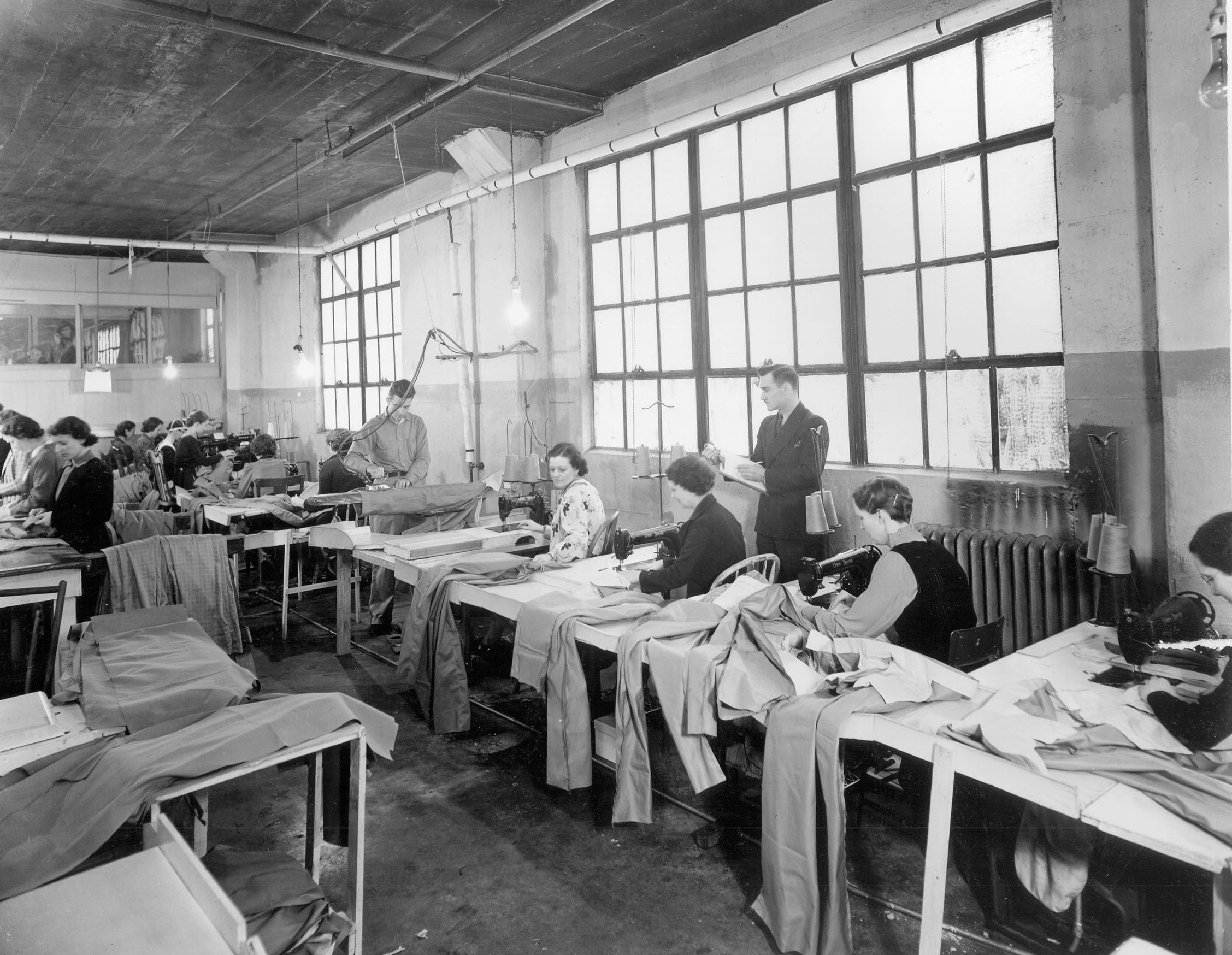 A black and white picture of people working at tables sewing pants