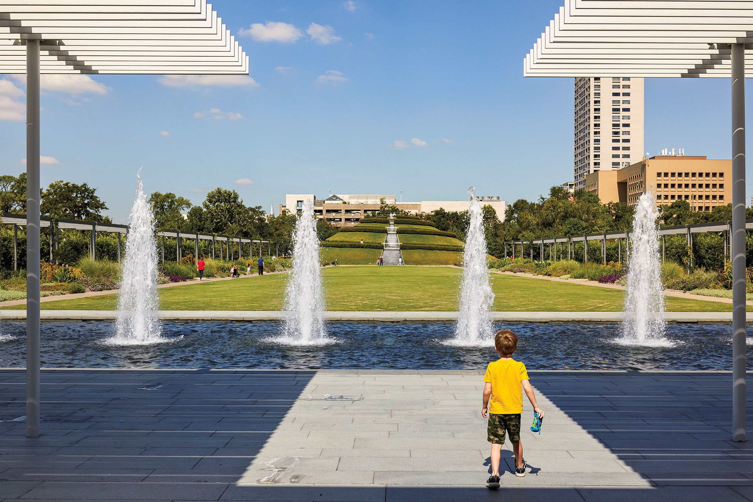 A young man in a yellow shirt stands in front of a large blue fountain, with green grass 