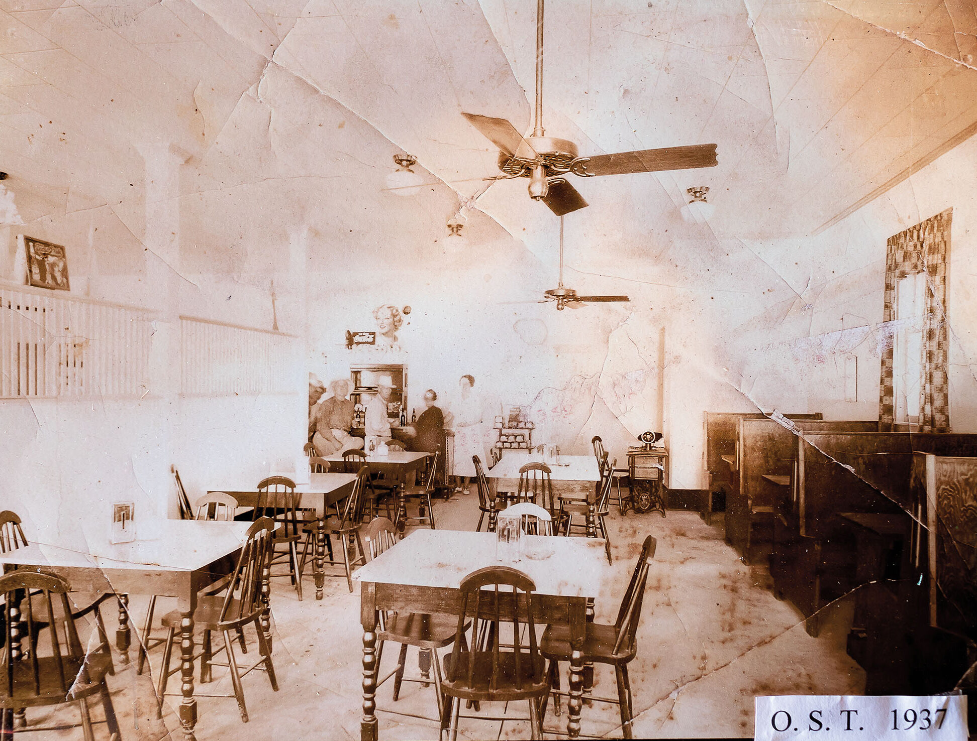 A sepia-tone historic photograph of the inside of a restaurant with ceiling fans, tables and chairs