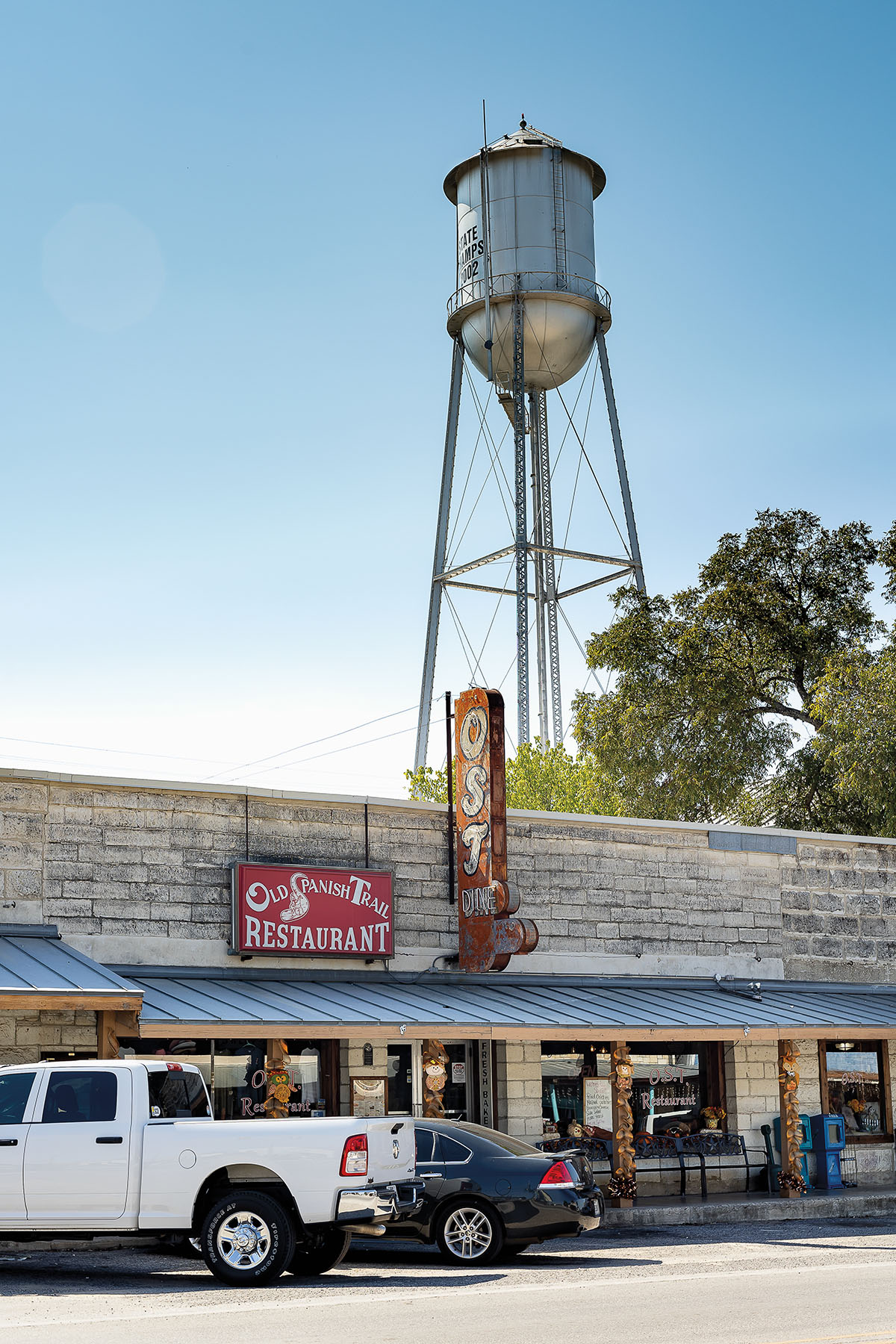 A tall silver water tower over a white limestone building with trucks parked out front