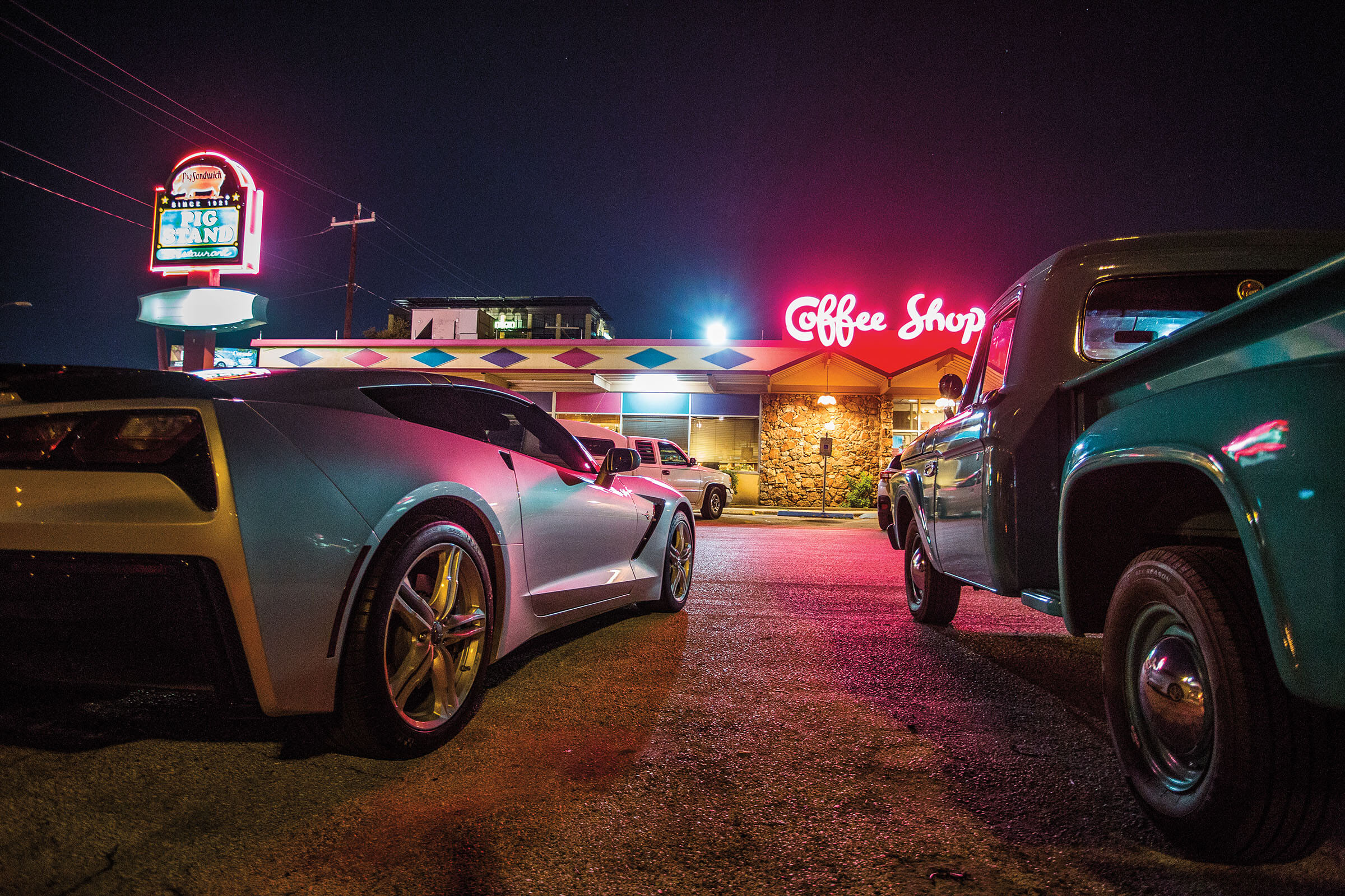 Bright pink neon reading "Coffee shop" illuminates the area behind a group of parked cars