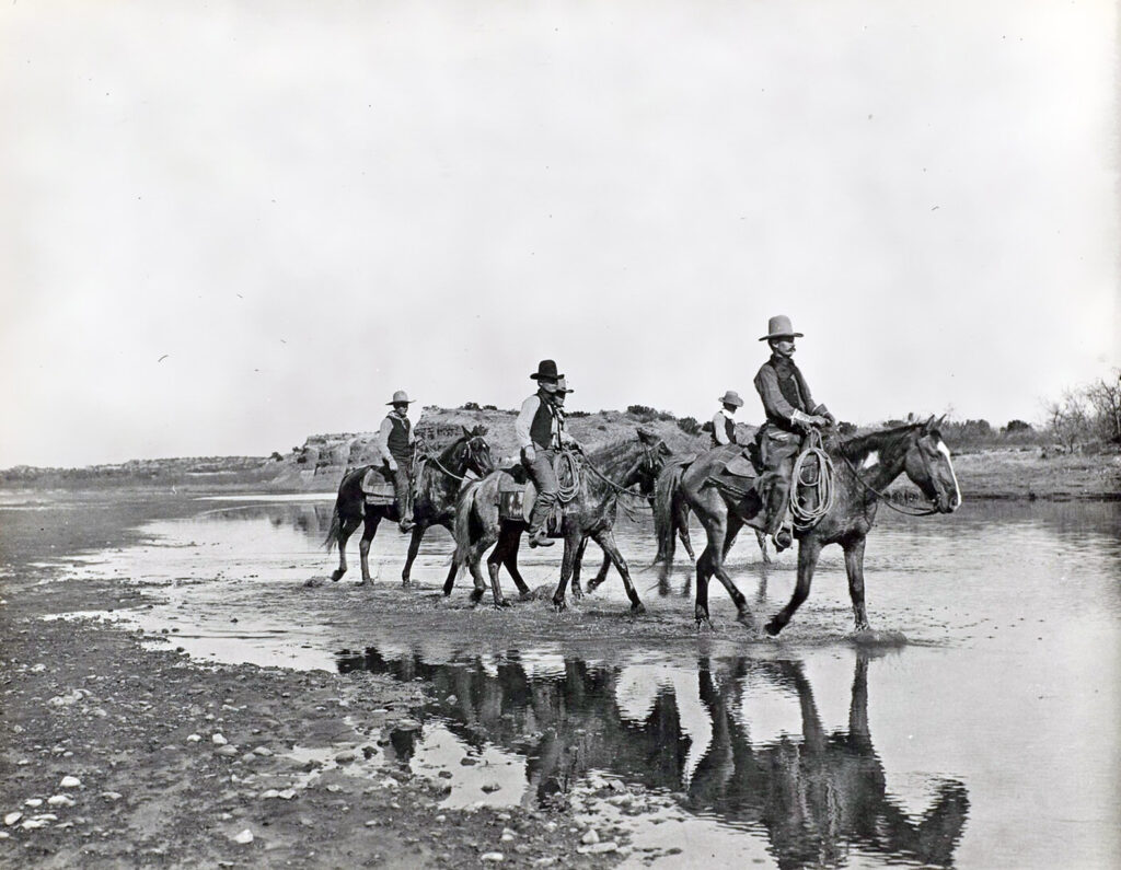 Cowboys on horseback ride through a small creek.