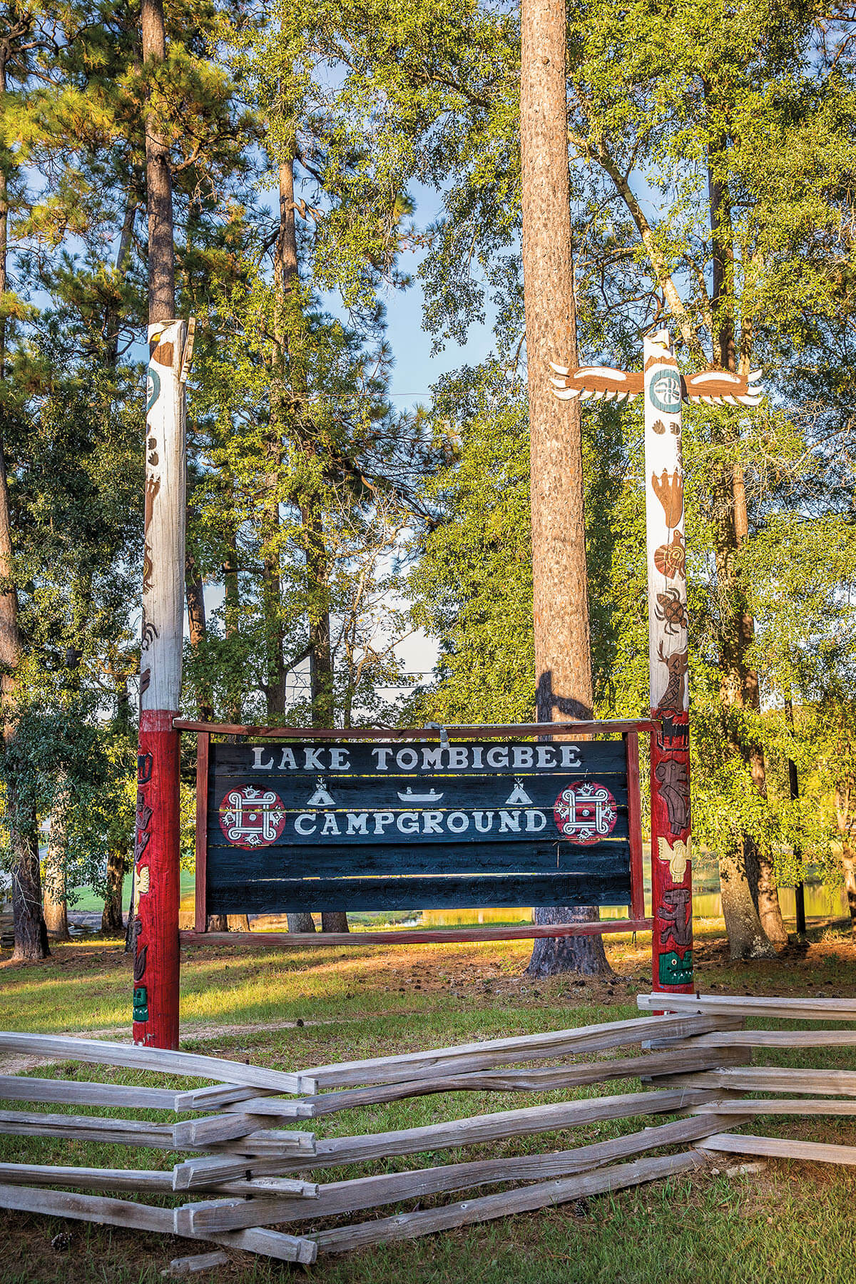 A wooden sign reading "Lake Tombigbee Campground" next to two large totem poles in a tall pine forest