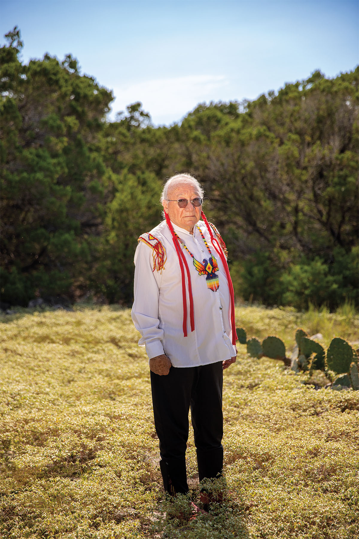 A man in traditional Native American wear, a colorful necklace and long red braids, stands in a field with prickly pear cactus