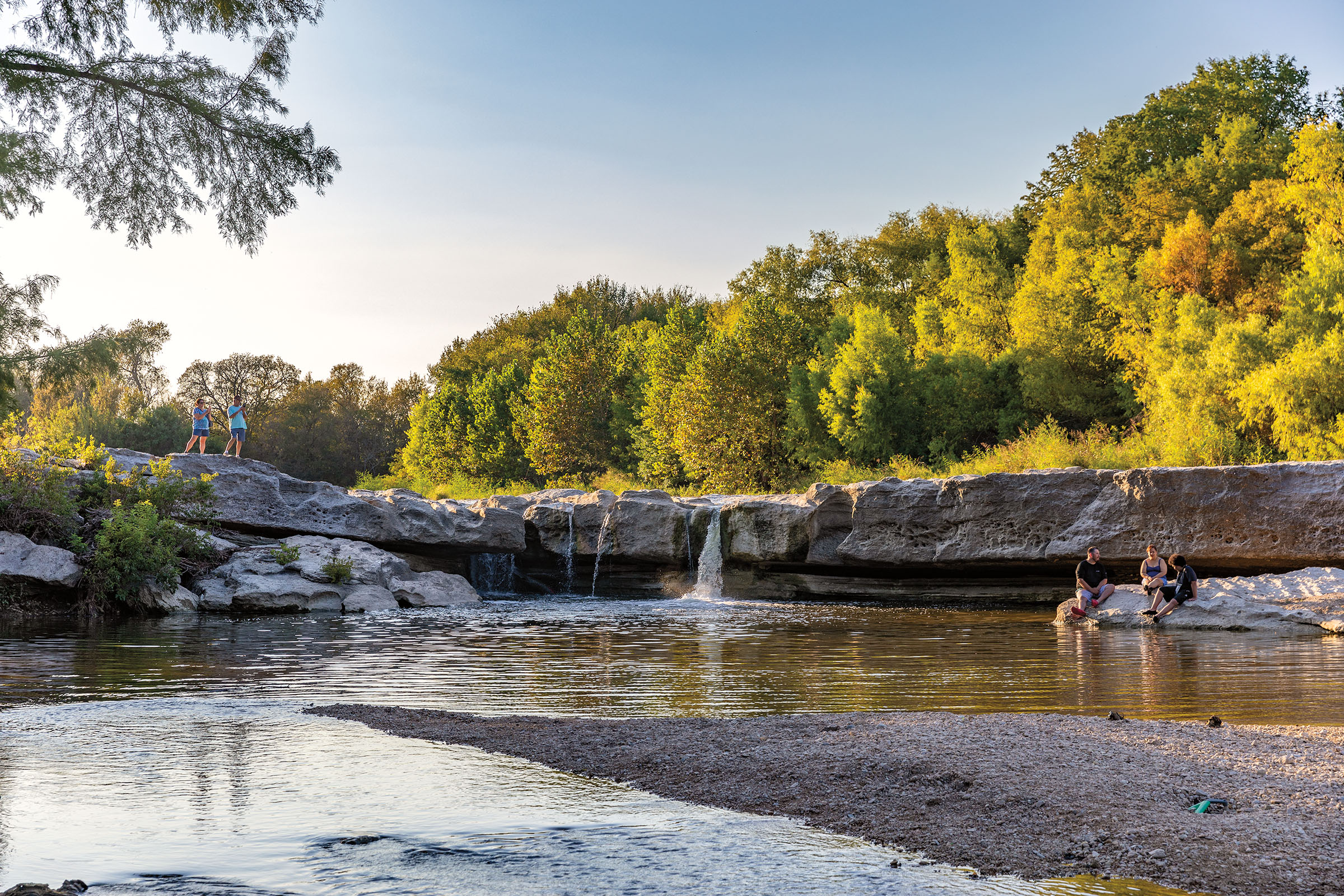 A view of green trees over a small waterfall in limestone rocks pooling into a large body of water. people stand and wade in the water.