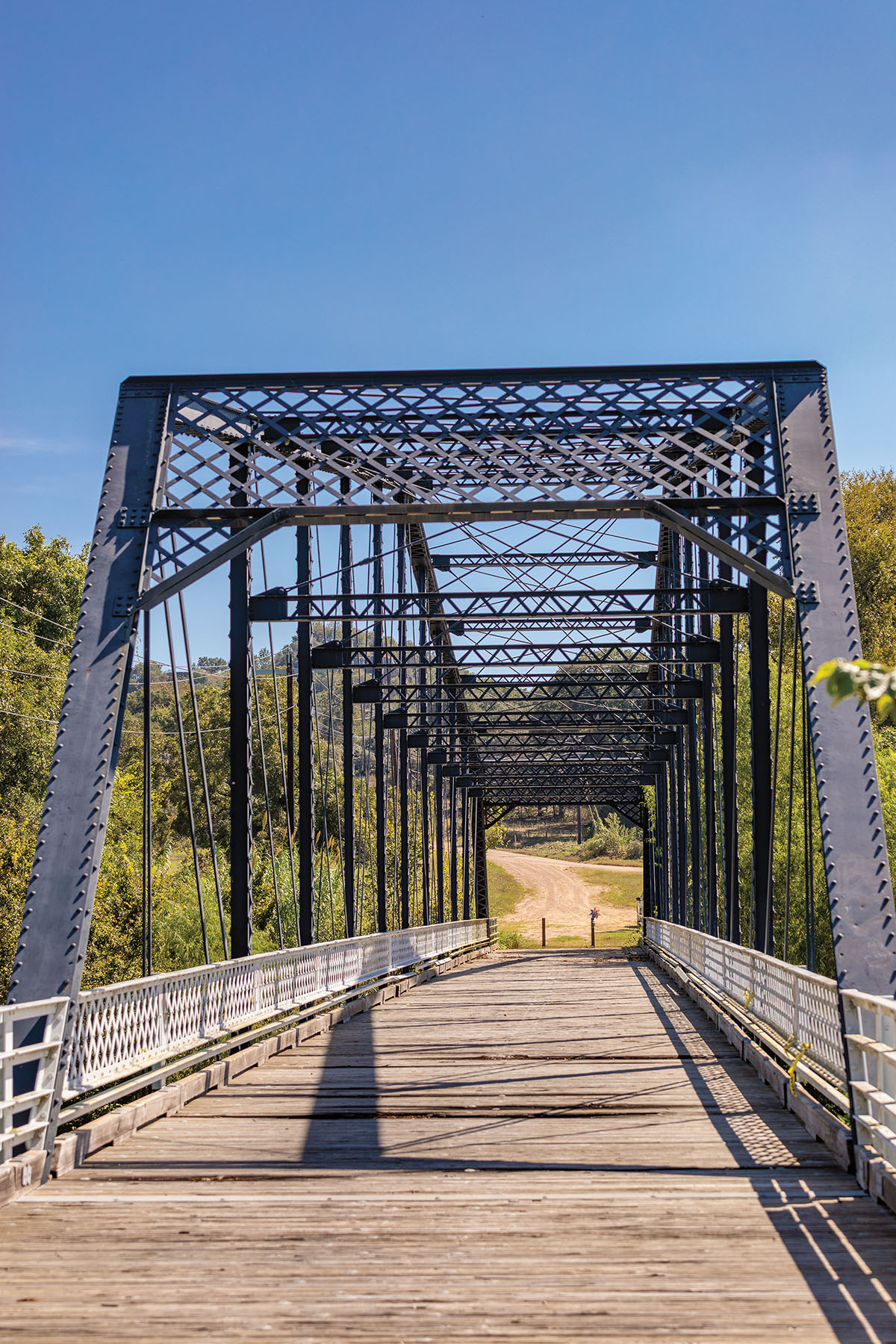 A gray metal bridge with a wooden road base connected to a dirt road surrounded by trees