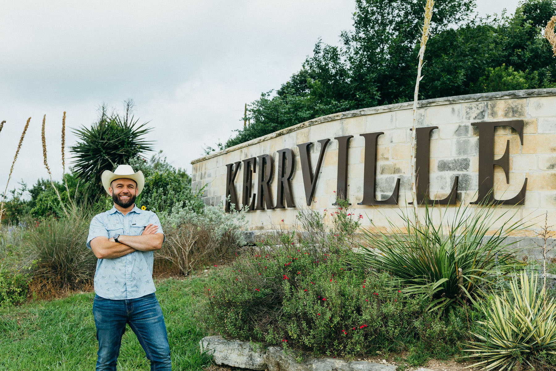 A man in a cowboy hat stands next to a stone sign reading "Kerrville"