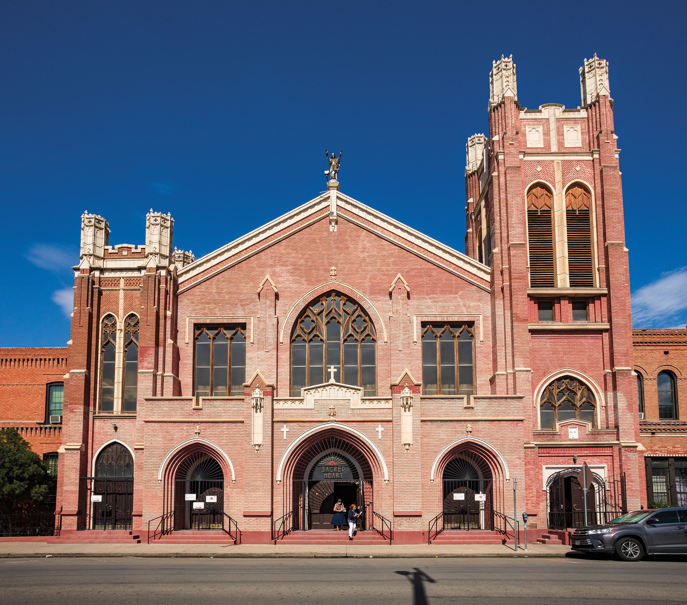 The red brick exterior of a church with a large steeple on the right side, shorter one on the left side, and two people standing out front