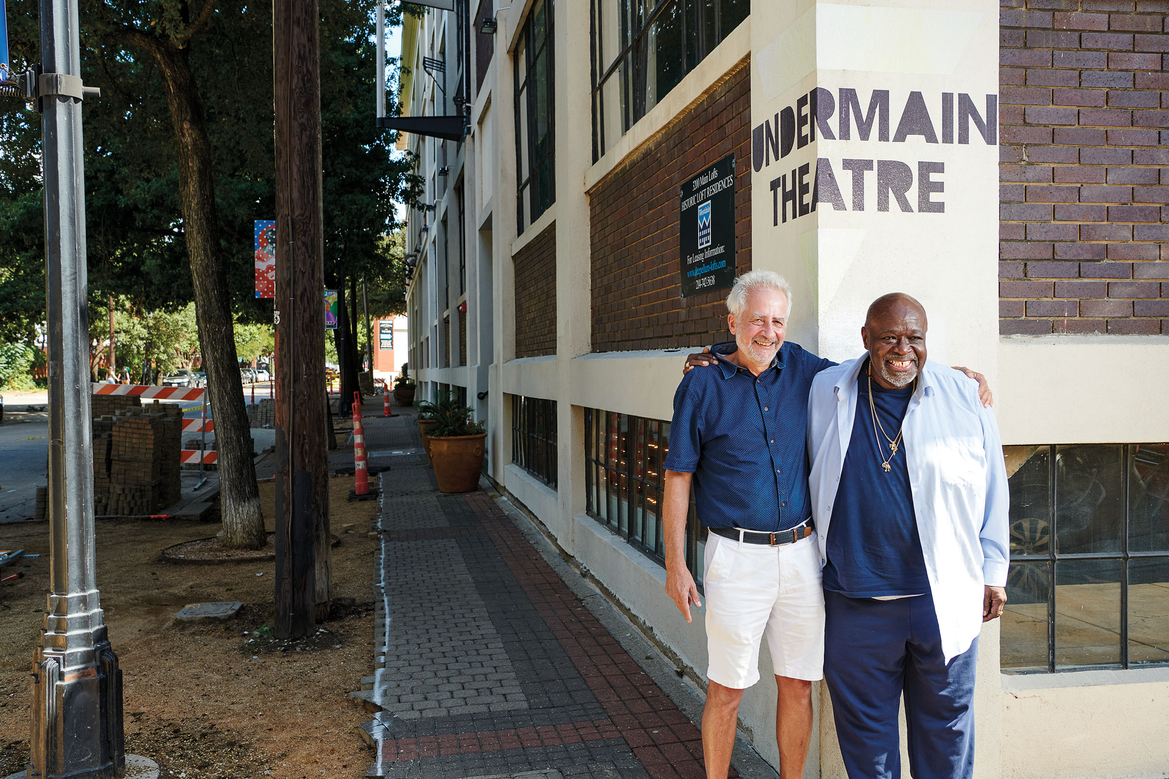 Two men stand with their arms around each other in front of a white and brick building