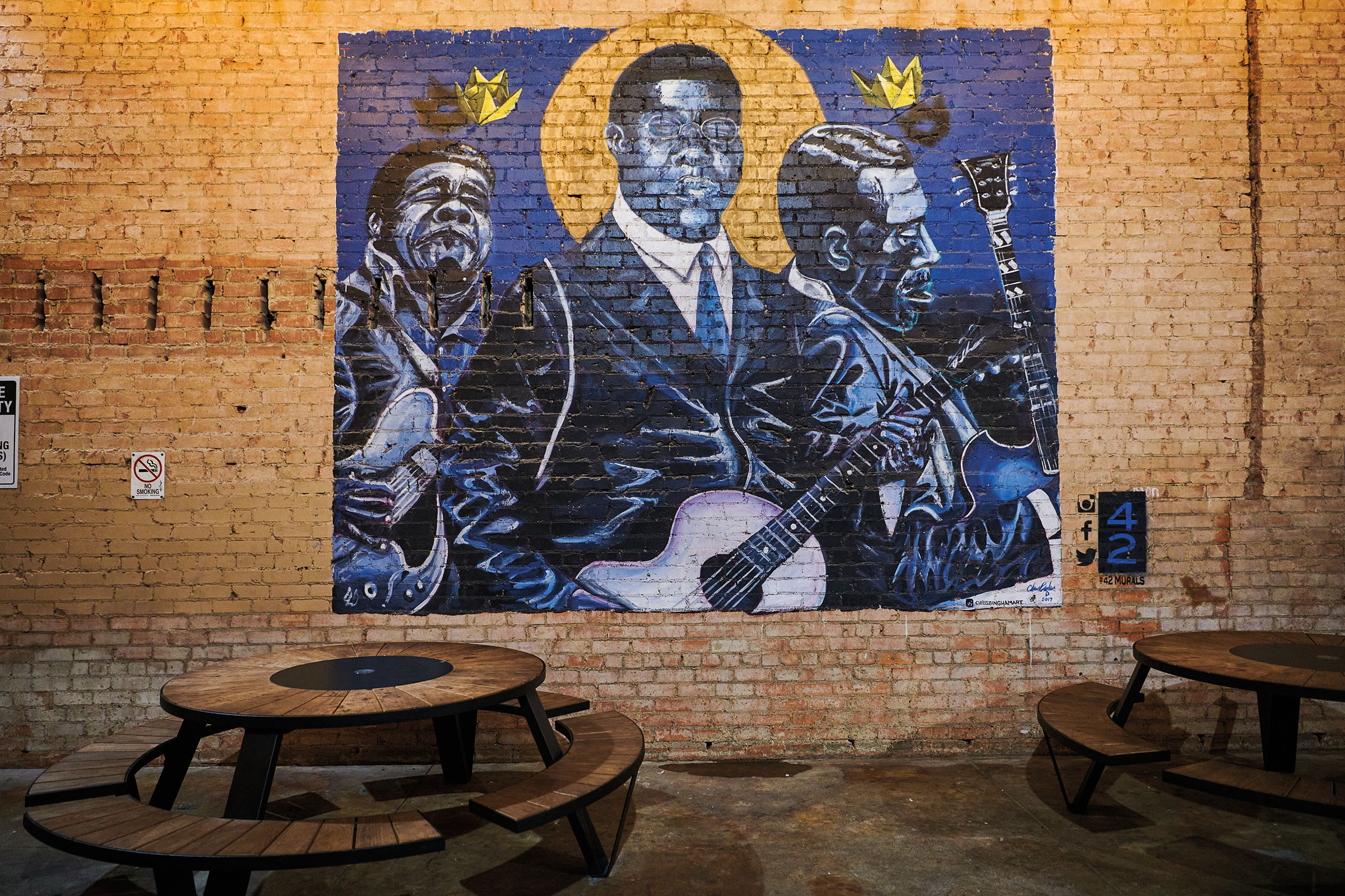 A dark blue mural painted on yellow bricks in front of brown wood picnic tables