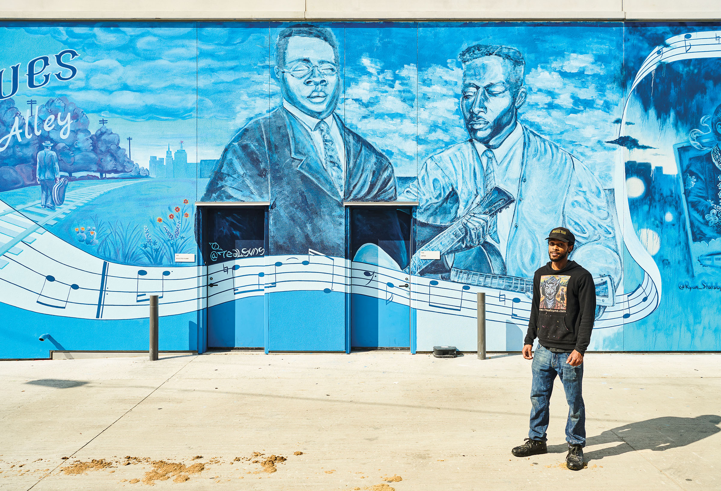 A man in a black sweatshirt and jeans stands in front of a blue-toned mural with two men and musical notes