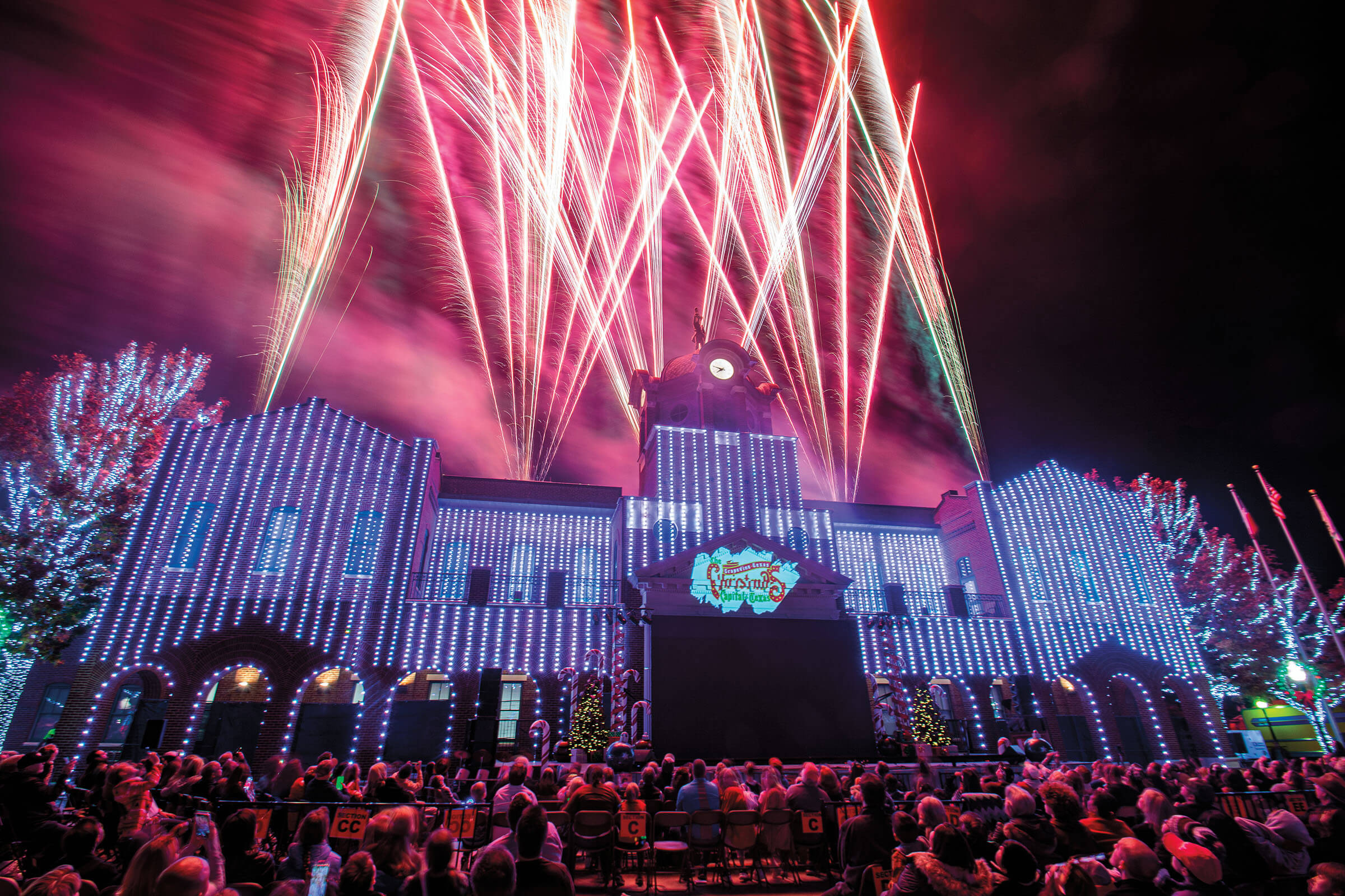 Bright red fireworks illuminate the back of a purple-white building with a crowd of people in front