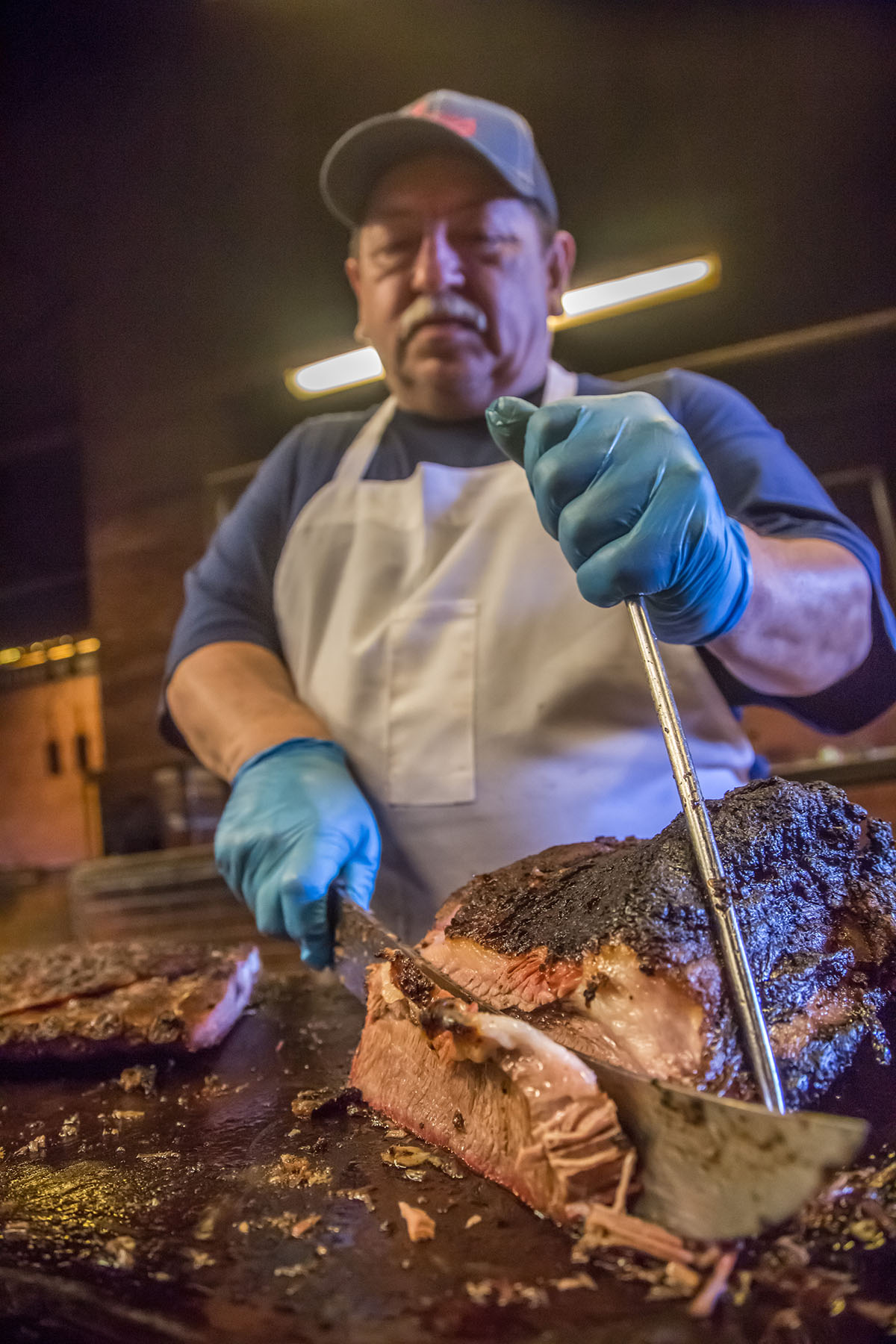 A man in a baseball cap holds a knife and steel to slice a fatty piece of brisket with dark bark.