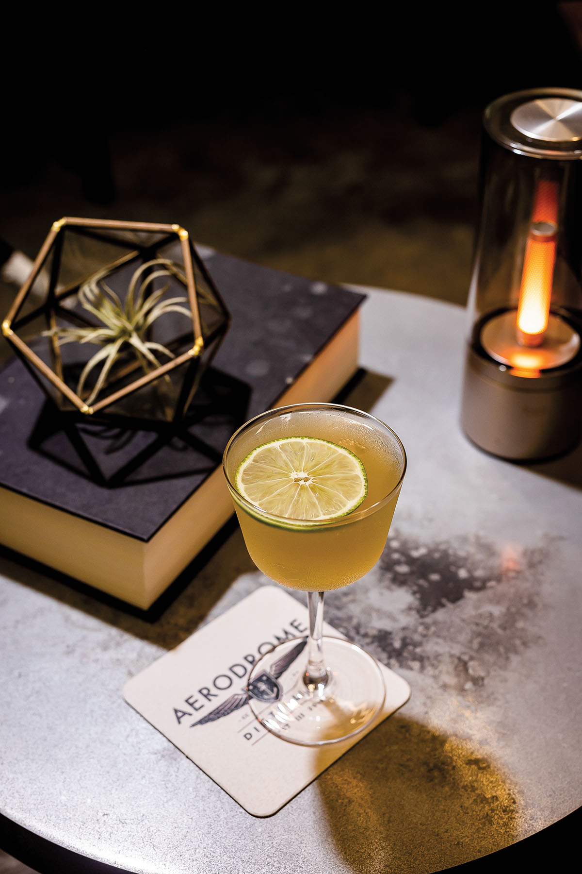 An elegantly-presented cocktail in a tall glass with a slice of lime on a coaster reading "Aerodrome"