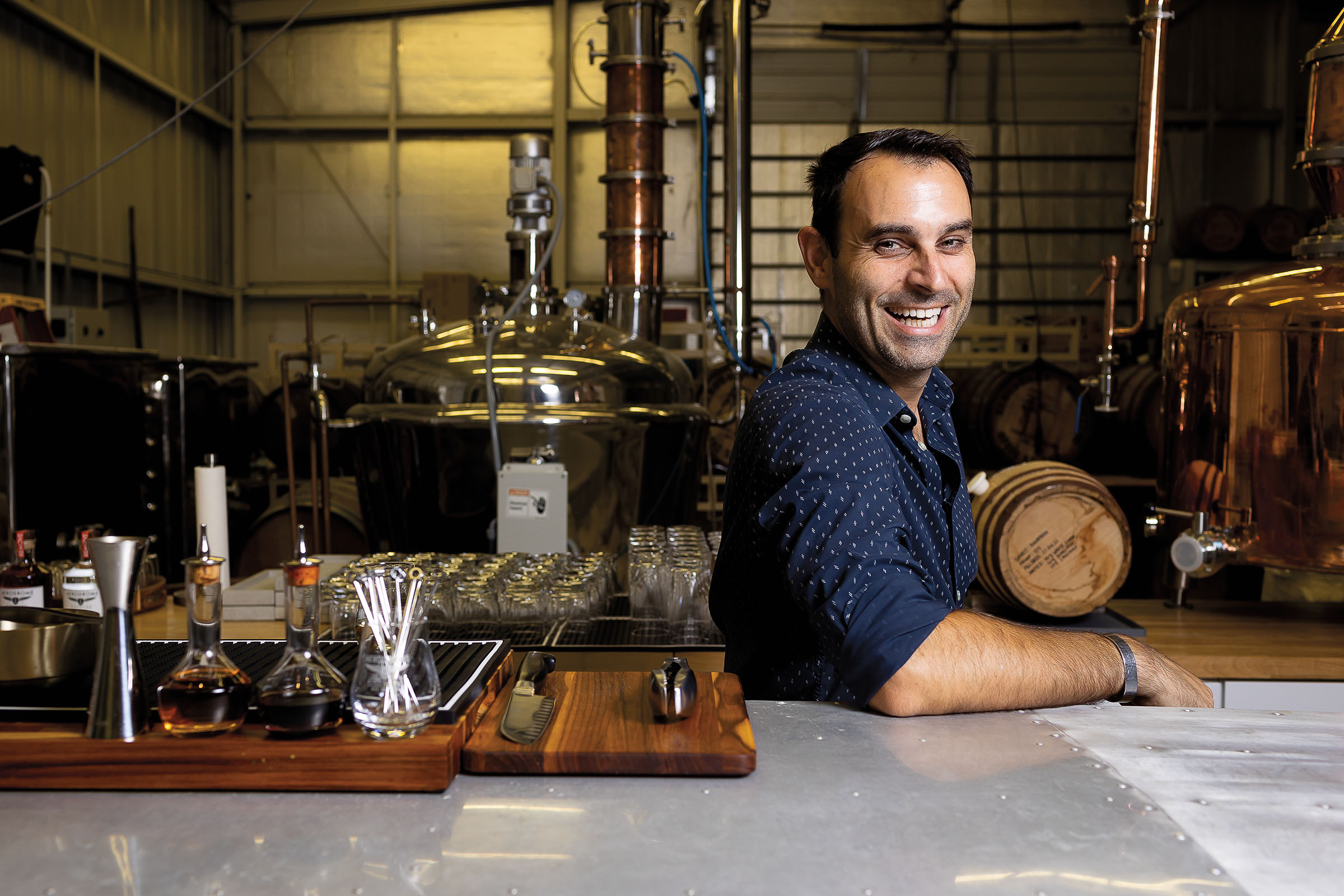 A man in a patterned blue collared shirt laughs with his back to glass and metal distilling equipment