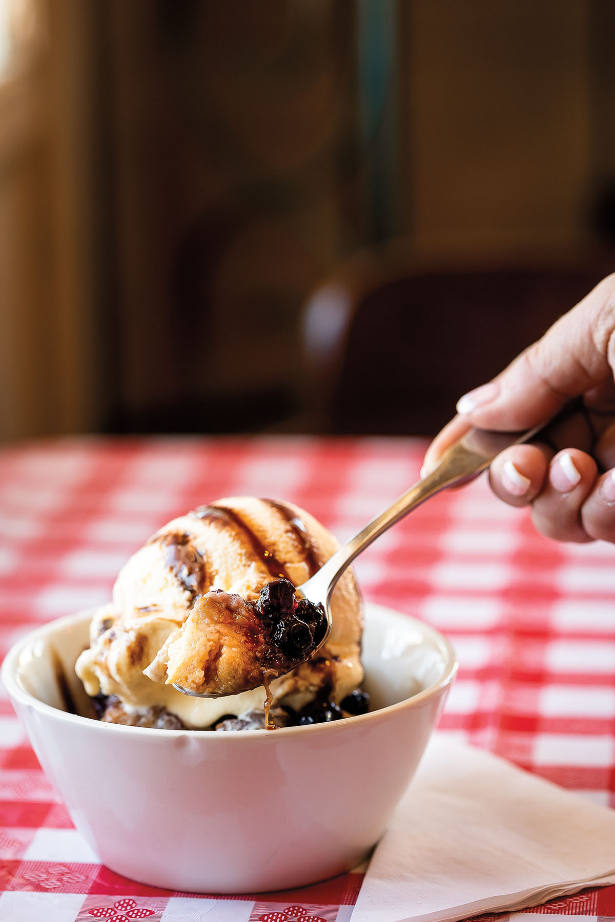 A spoon dips into a white bowl filled with golden bread pudding topped with vanilla ice cream