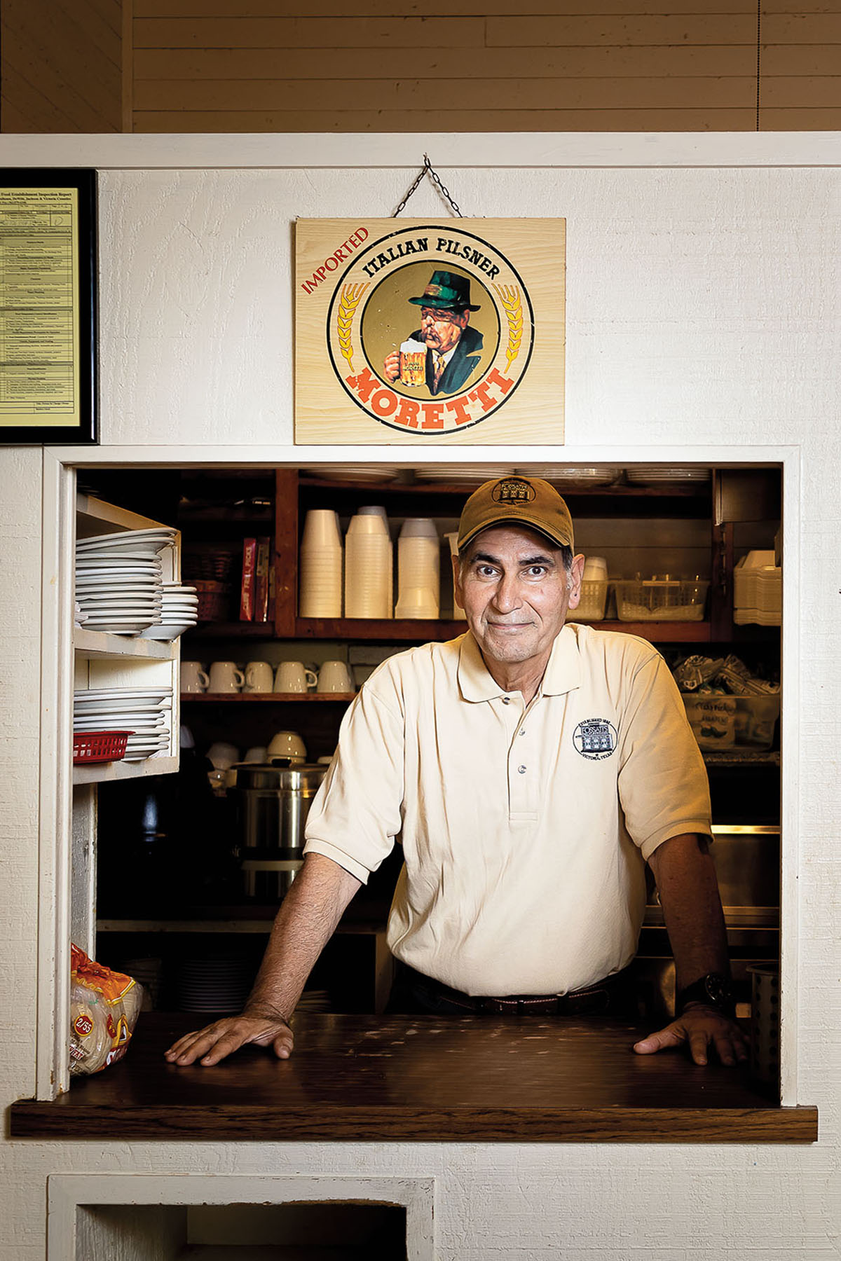 A man in a brown baseball cap stands in front of a window under a sign reading "Italian Pilsner - Moretti"