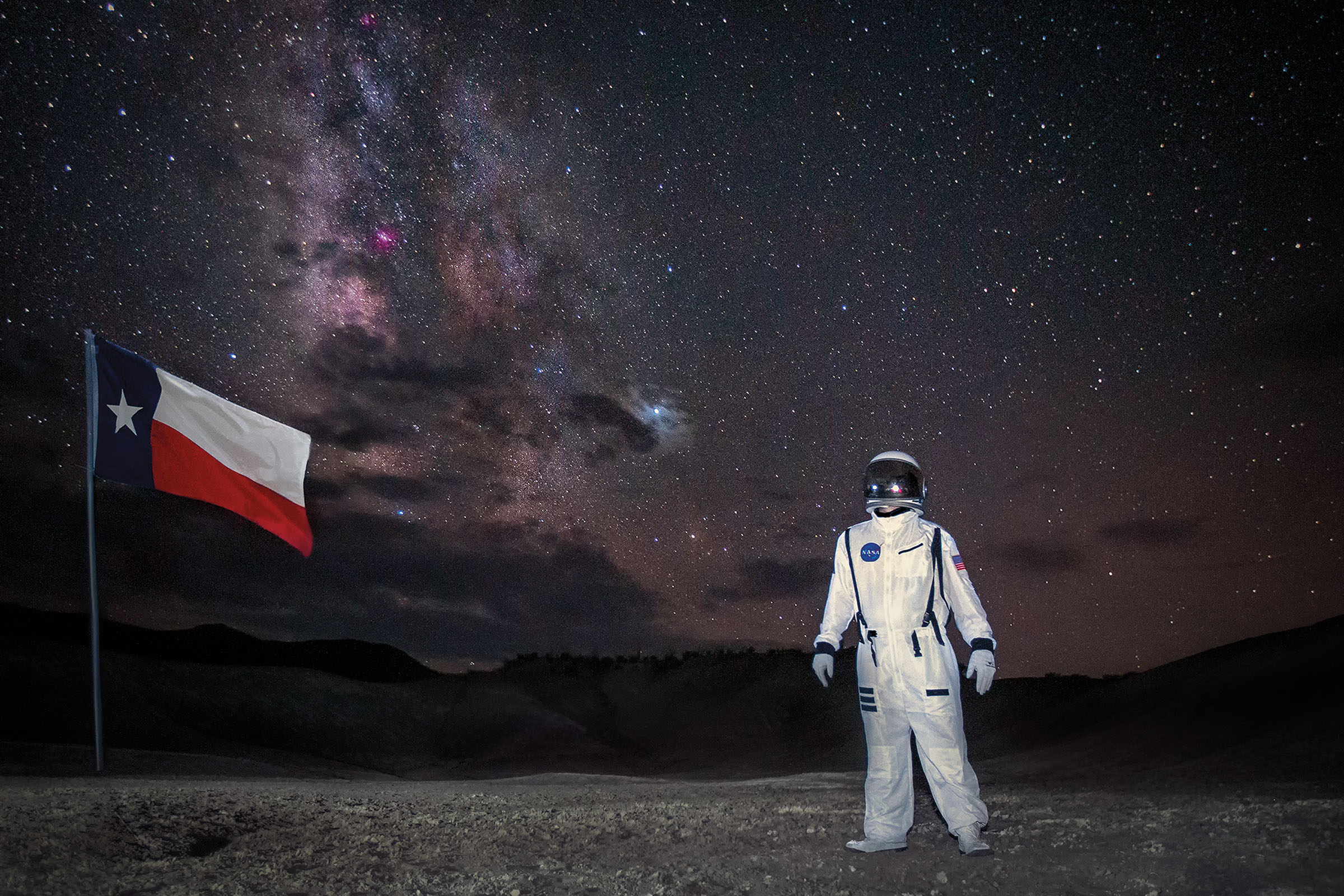A person in an astronaut suit stands next to a Texas flag underneath a large purple and black night sky filled with stars
