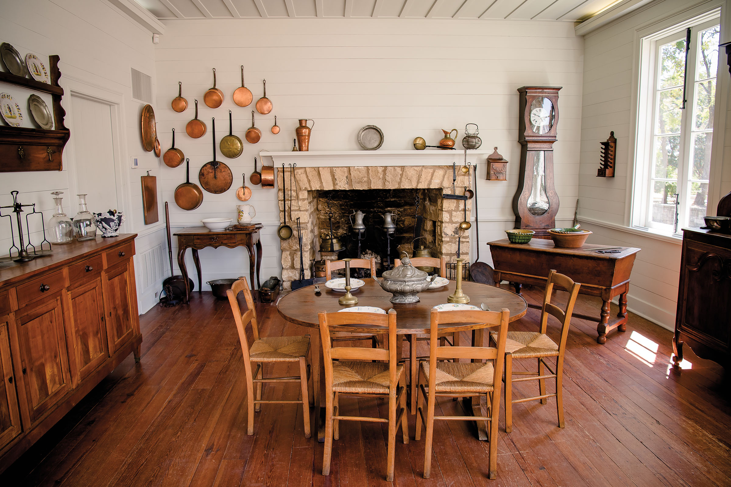 A period-era interior including white walls, copper pots, a large stone fireplace, and a circular wooden dining table with large brass candlesticks