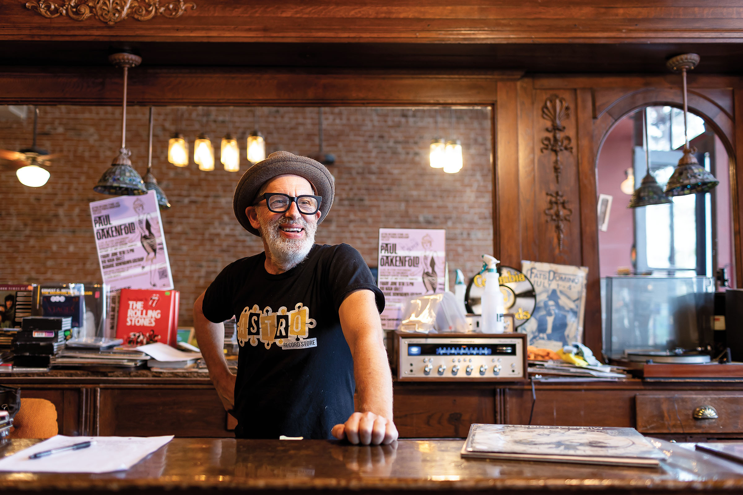 A man in a small brimmed hat and a blue t-shirt reading "Astro" stands in front of a brick wall and a vintage amplifier