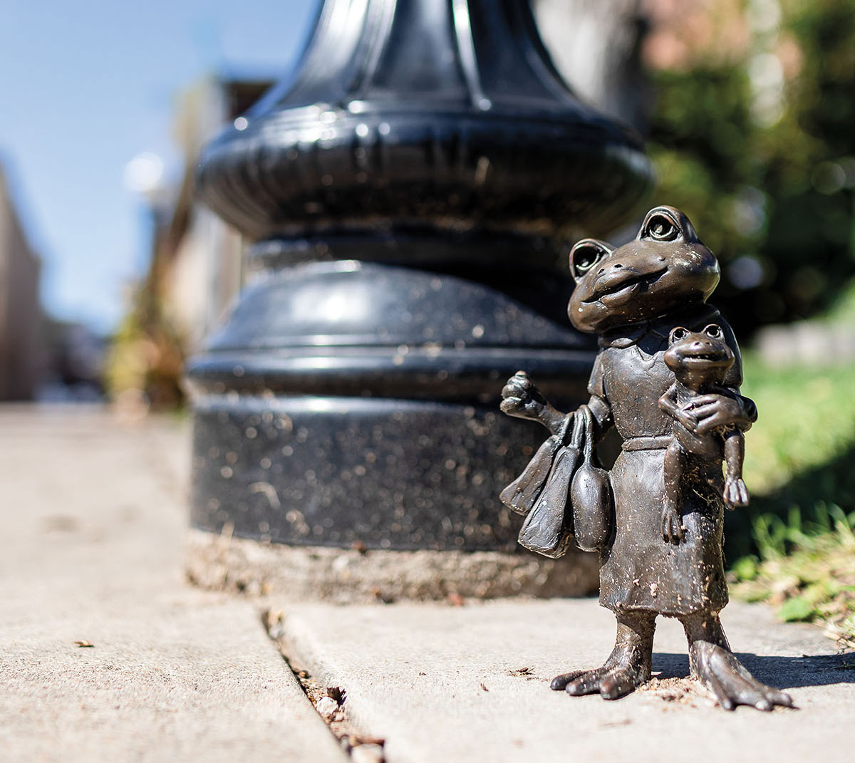 A bronze sculpture of a frog holding a smaller frog next to the base of a lightpost on a concrete slab