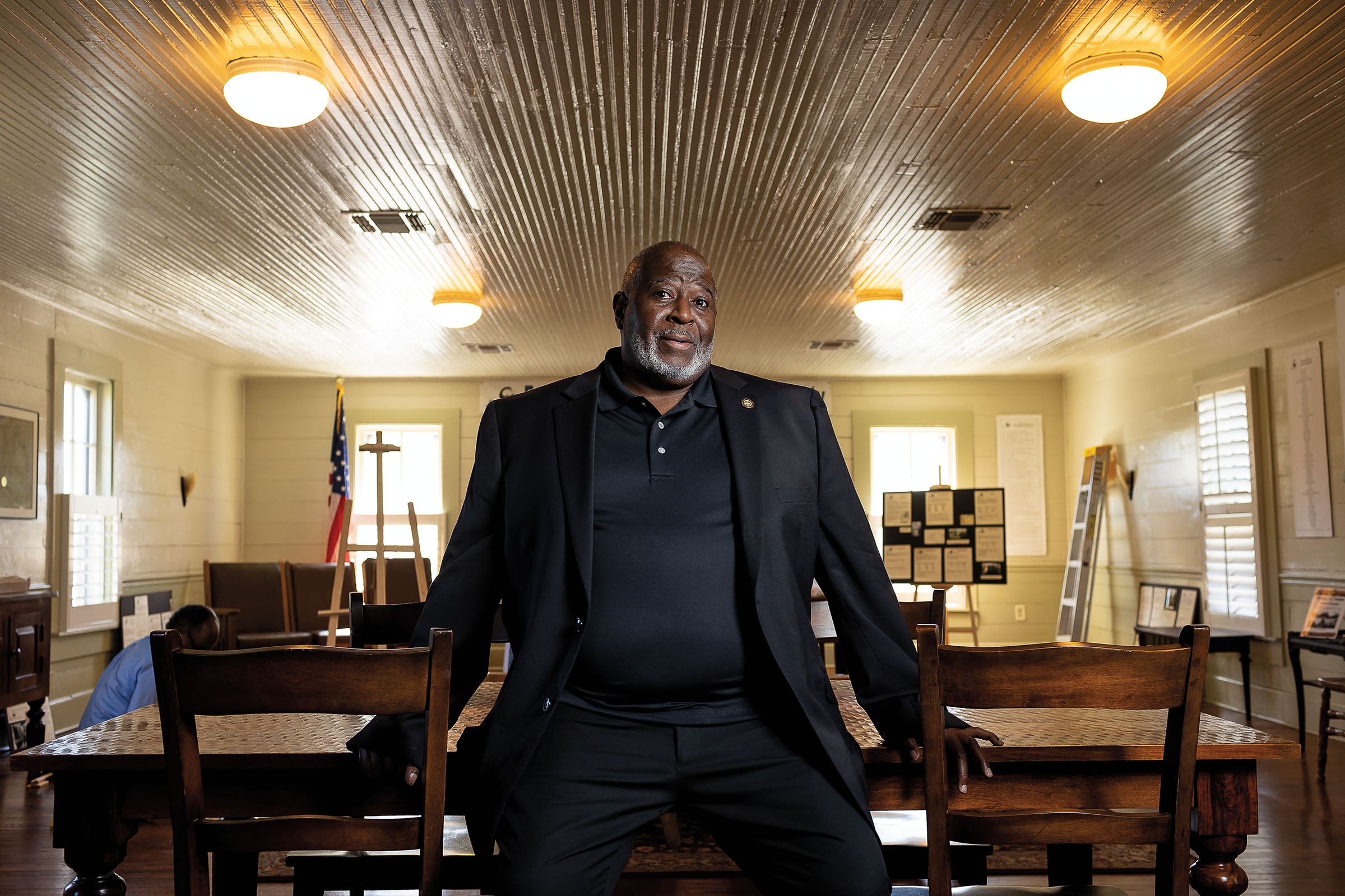 A man in a black polo shirt and black suit jacket sits on a wooden table next to wooden chairs inside of an old-fashioned room with overhead lighting and wood paneling