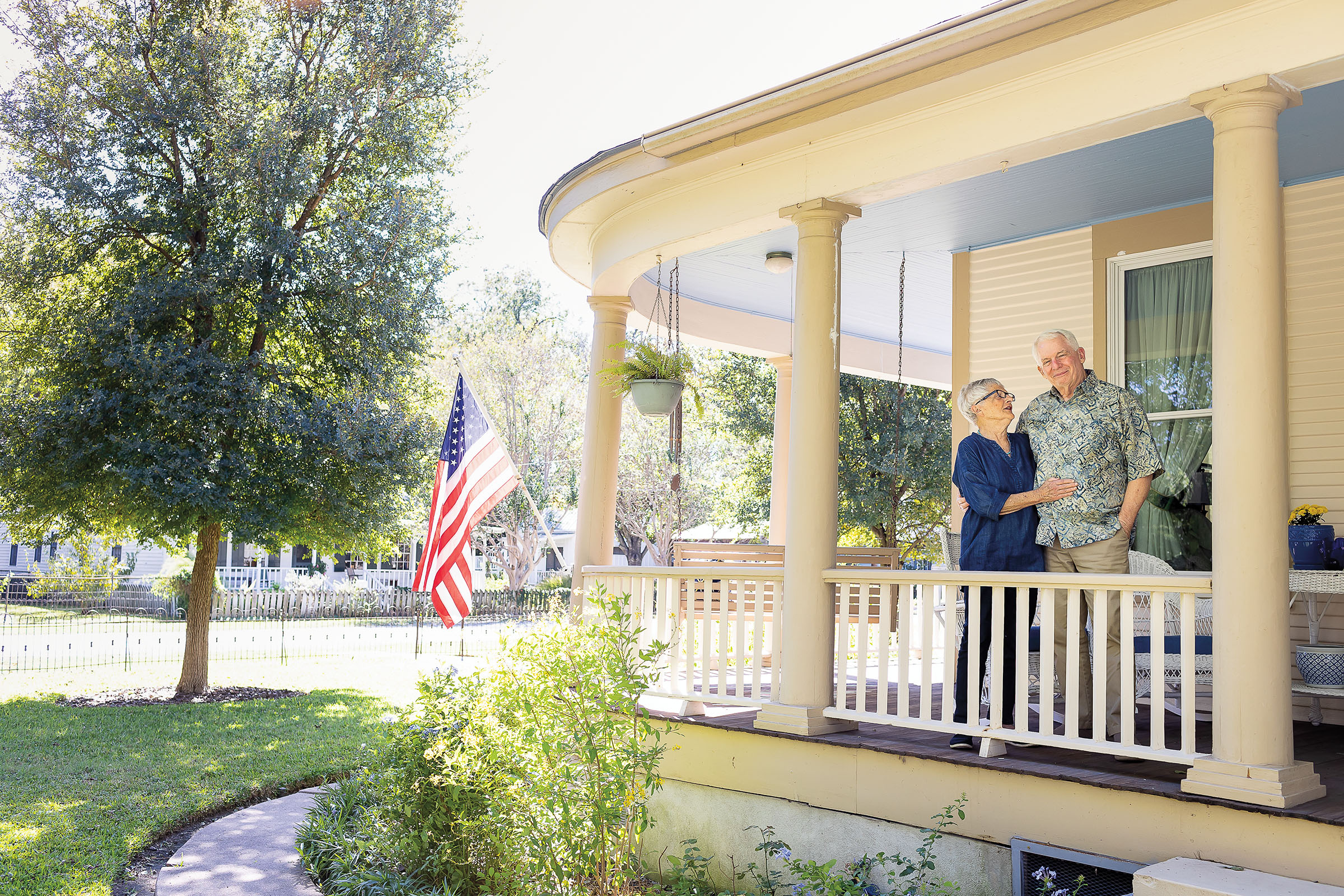An older woman and man stand on the porch of a large house with columns and an American flag on the porch