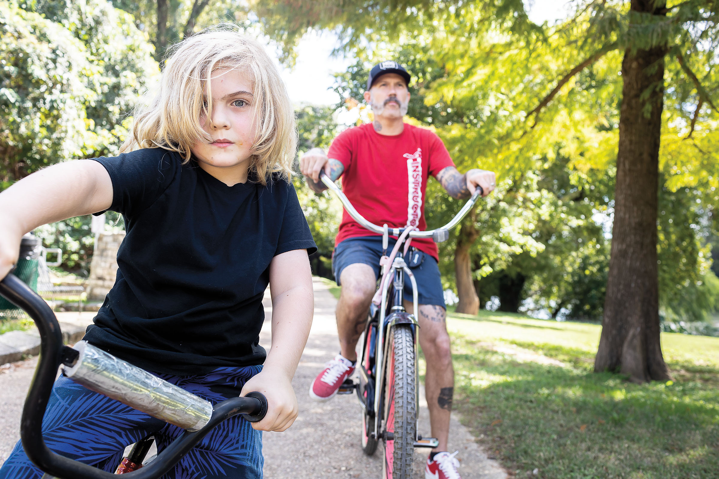 A young person with long blonde hair looks at the camera from their bike while a man on a larger bike in a red shirt looks off into the distance