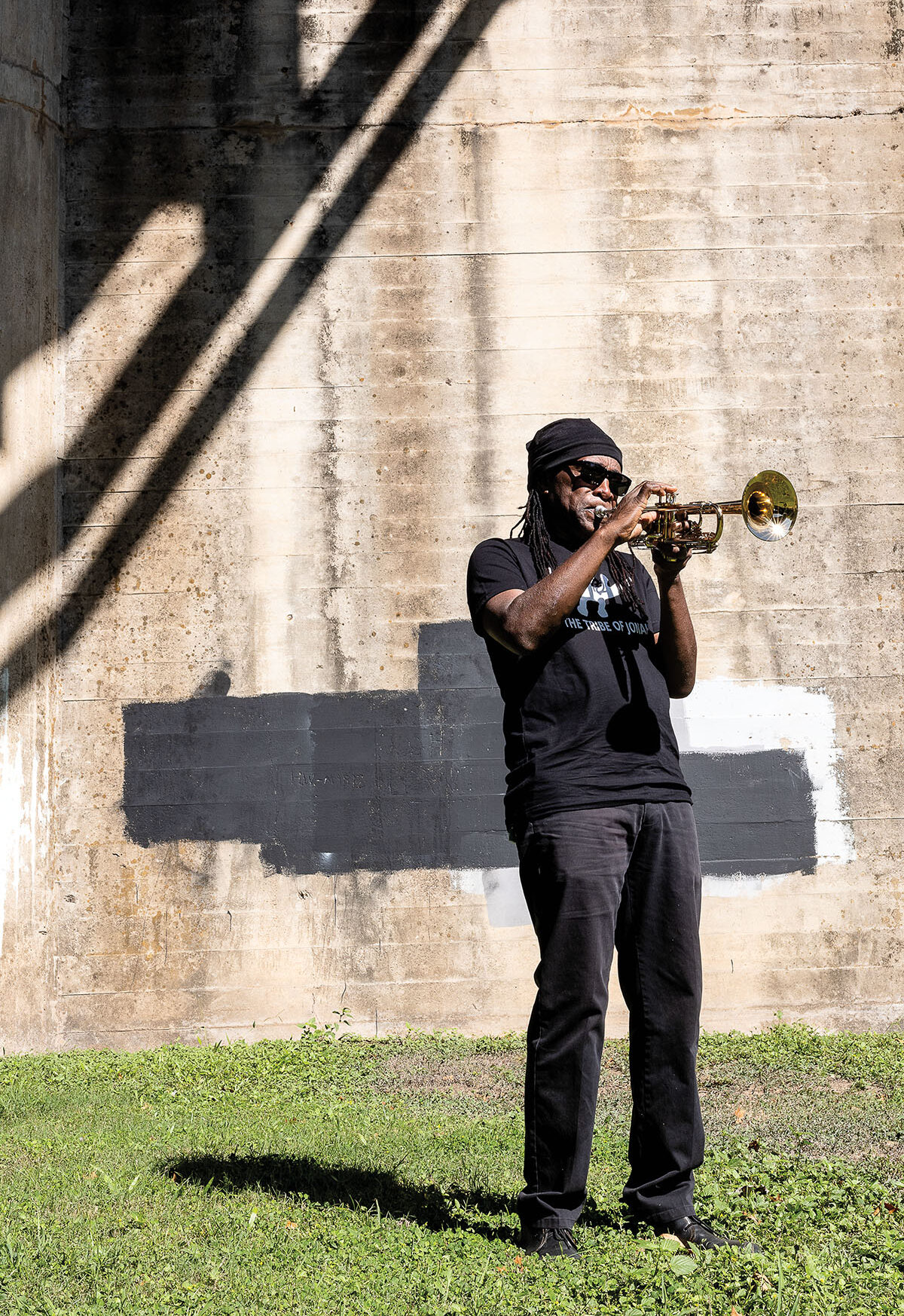 A man in a black hat and t-shirt plays a trumpet in front of a concrete background on grass