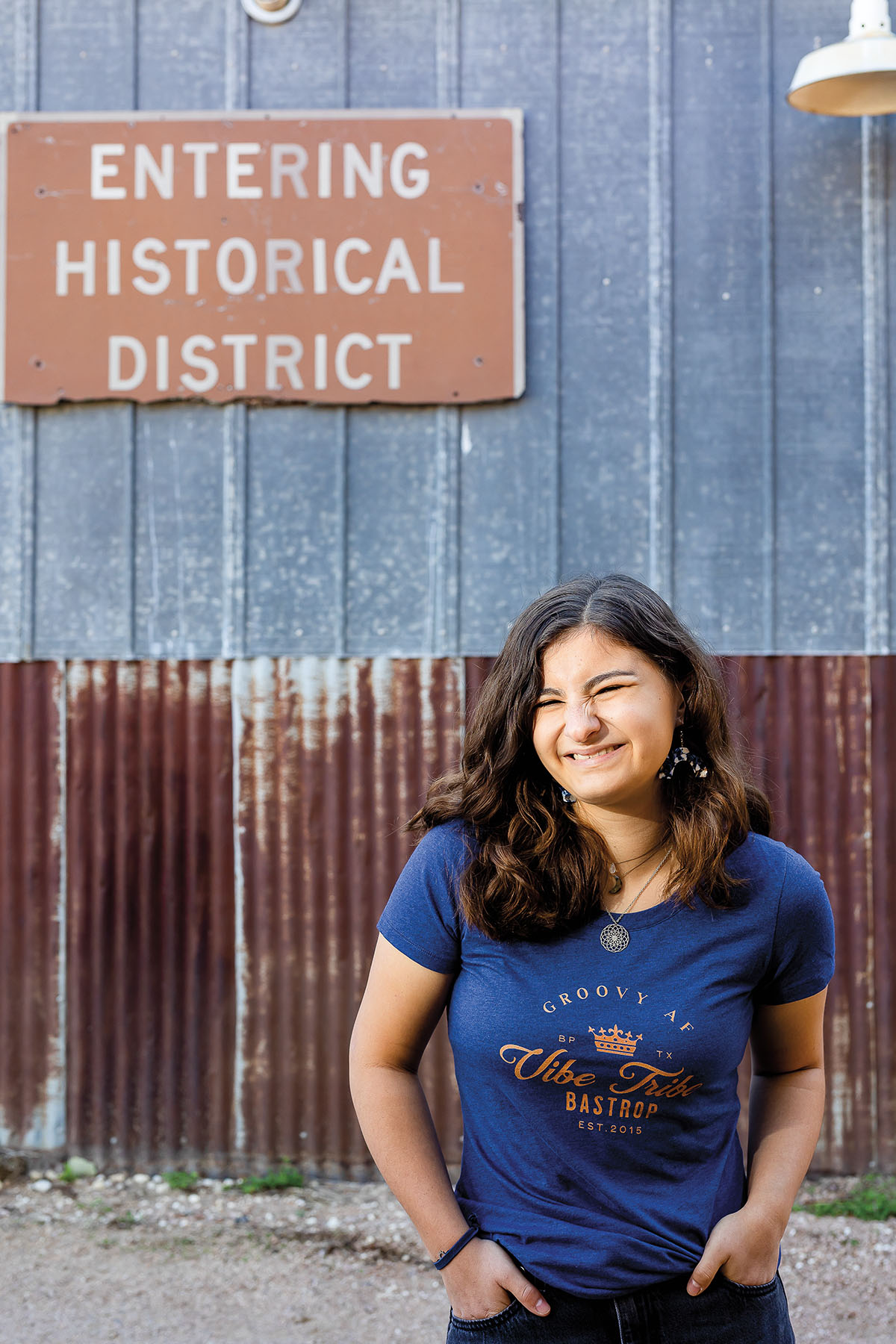 A woman in a blue shirt reading "Groovy at Vibe Tribe Bastrop" stands in front of a metal building with a brown sign reading "Entering Historical District"