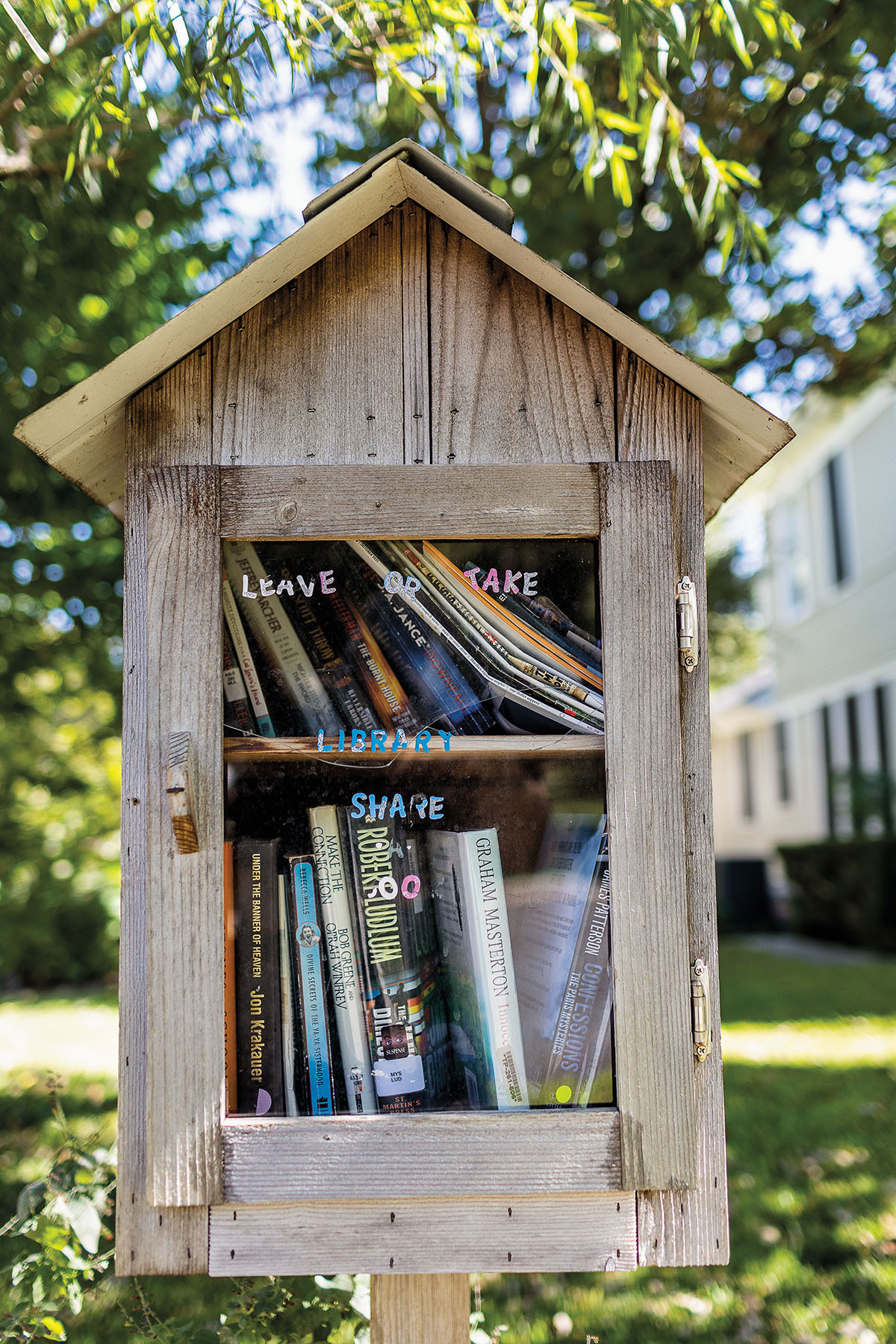 A small wooden house-shaped box with a clear door revealing a series of books and handwriting that reads "Leave Or Take Library Share"