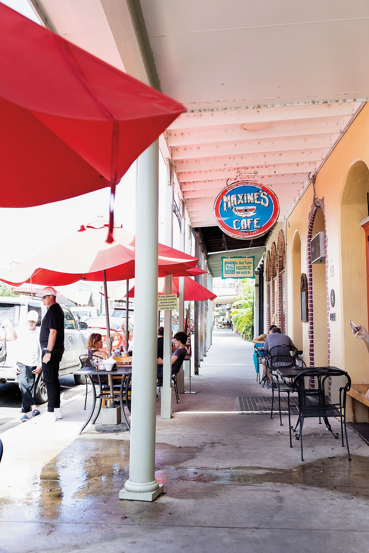 Red umbrellas line the sidewalk under a old neon sign reading "Maxine's Cafe"