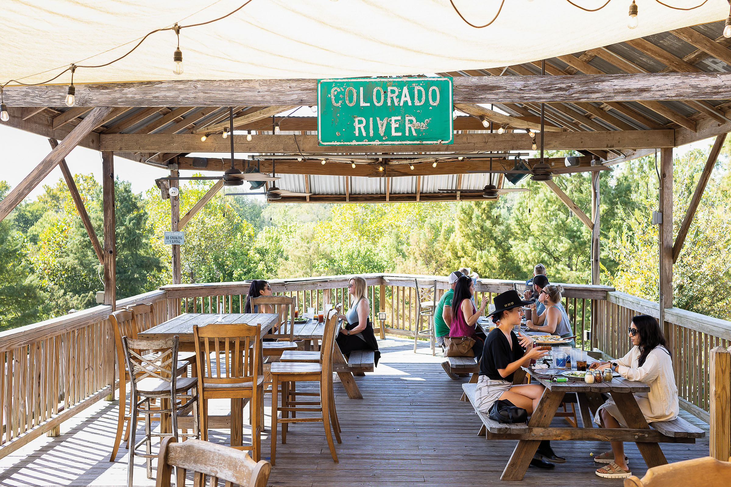 A group of people sit on a porch in a lush green setting under a green sign reading "Colorado River"