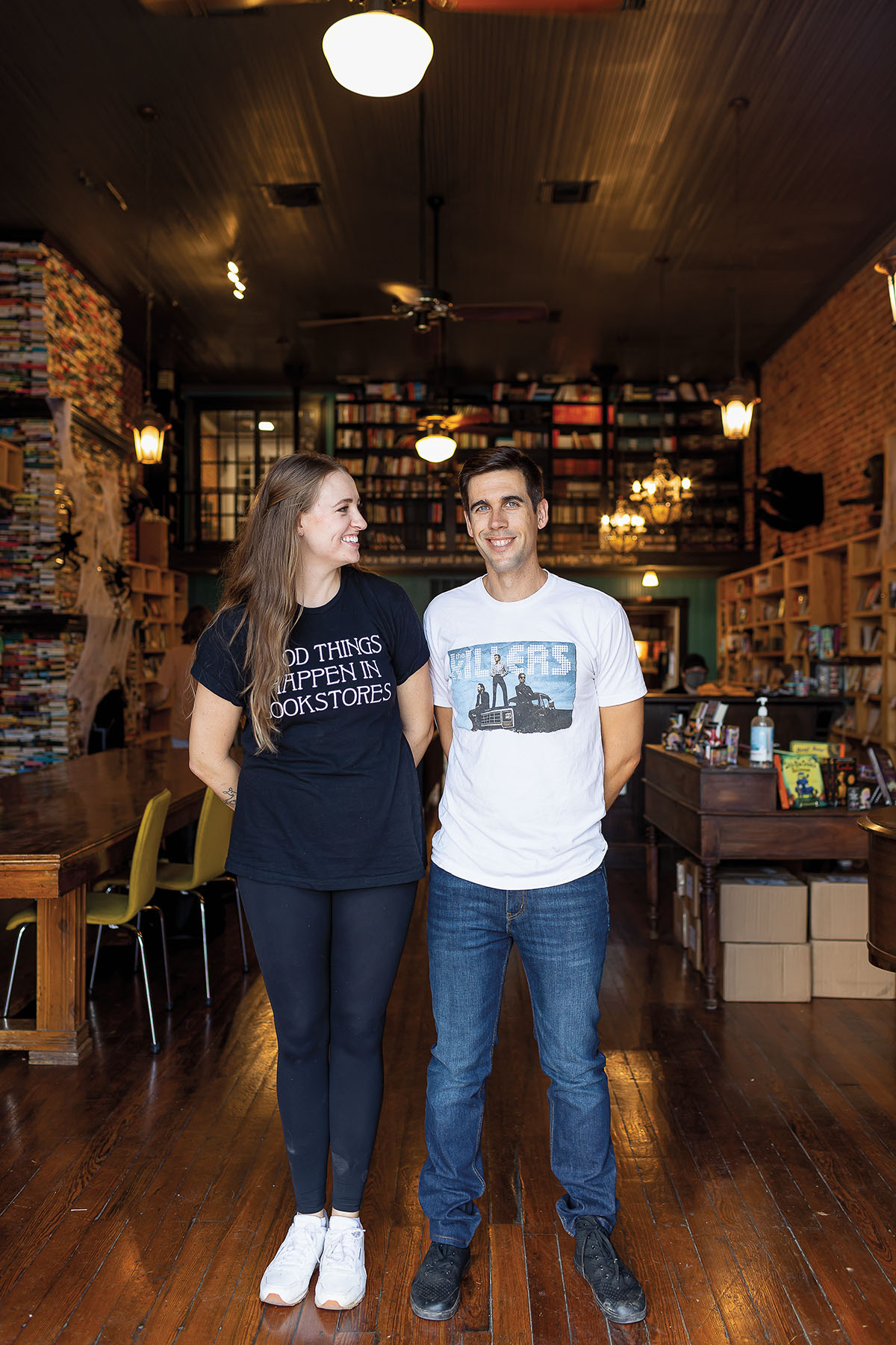 A woman and man, dressed in t-shirts and jeans, stand inside of a warm, wood, richly decorated room with books on shelves and a light overhead